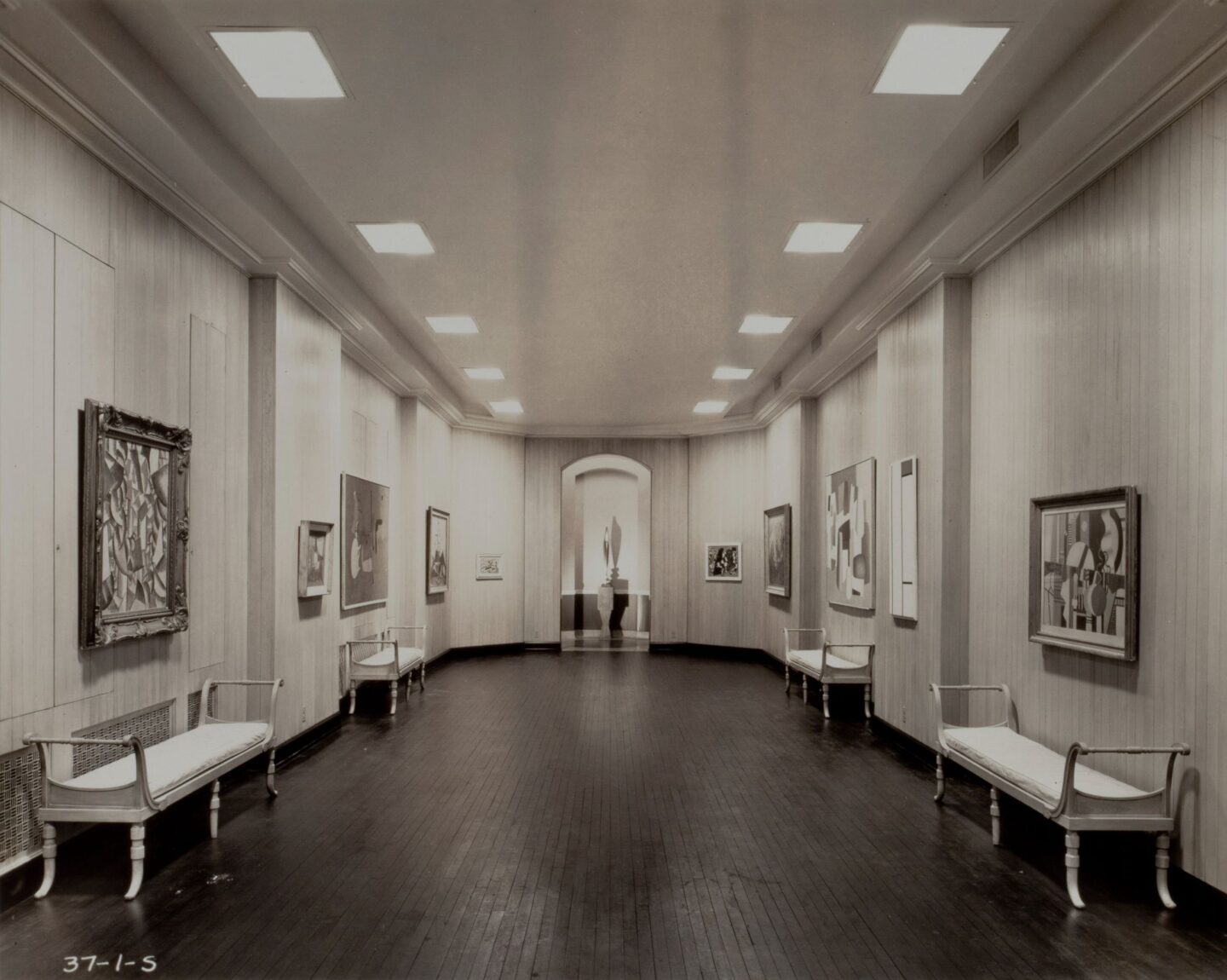 A large gallery interior. The left and right gallery walls are lined with five modernist paintings on each side. In the center and to the back of the gallery is an abstract golden sculpture of a bird.
