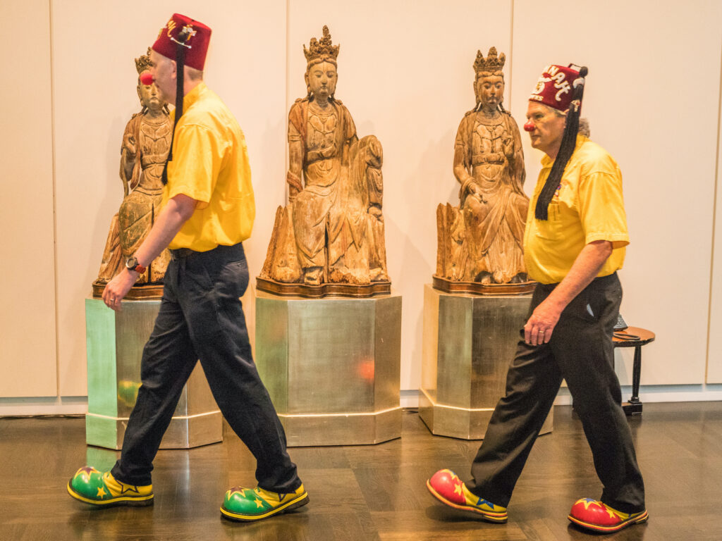 Two Shriners wearing a red clown nose and clown shoes walk past a series of ancient sculptures.