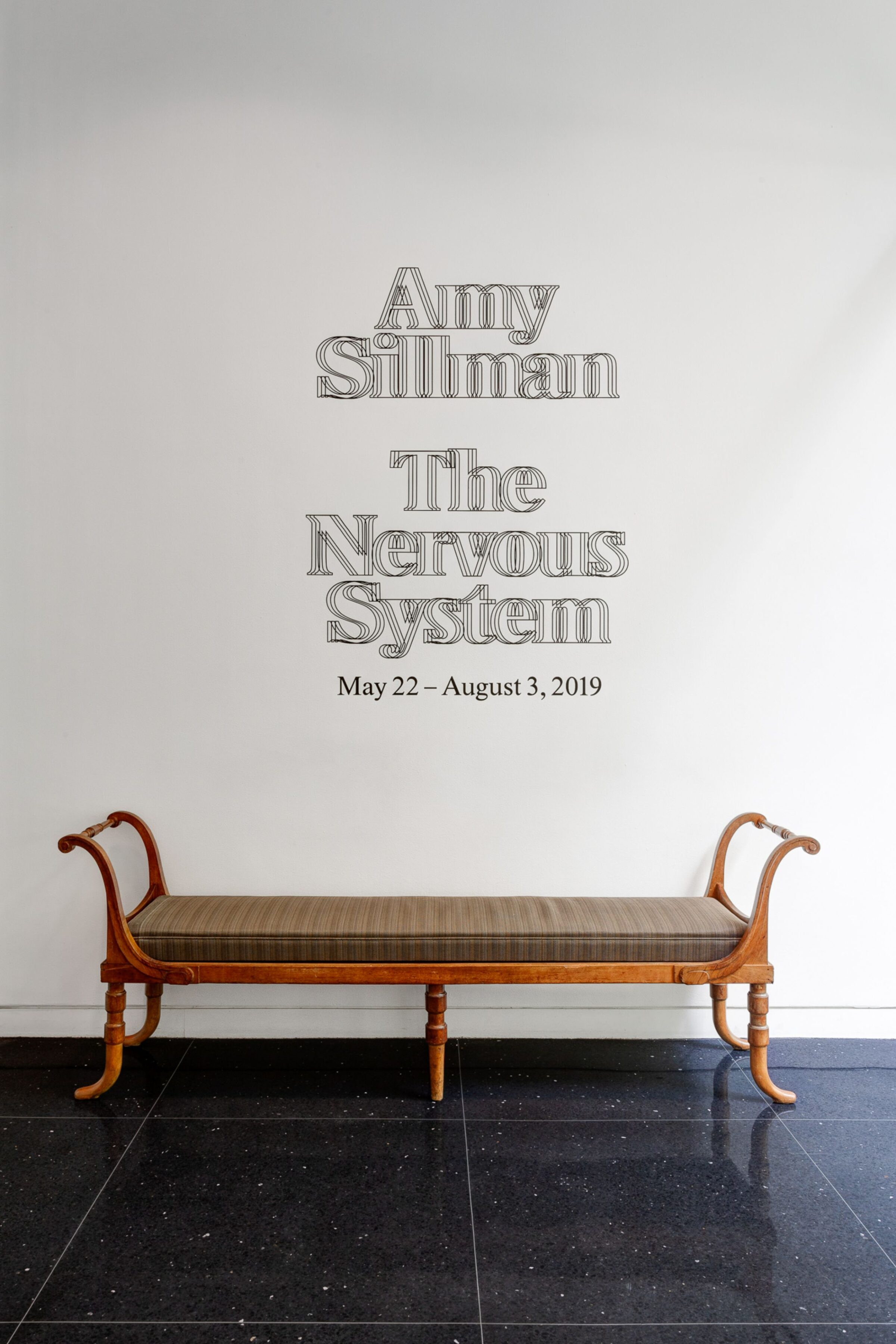 Gallery entrance of The Arts Club. Black text on a white wall reads, Amy Sillman: The Nervous System, May 22-August 3, 2019. In front of the wall sits a bench.