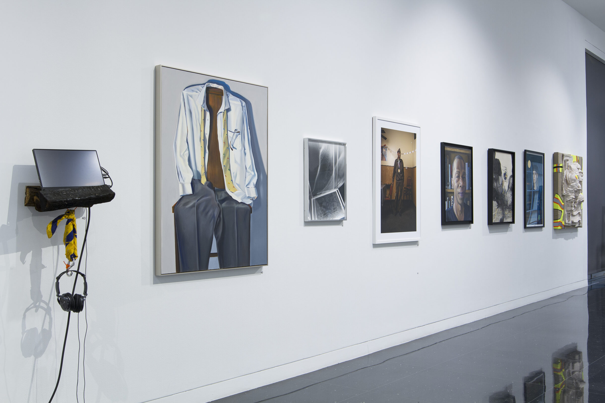 A white walled gallery space displaying a row of framed artworks. On the far left side is a makeshift audio and video installation next to a large painting of a men's suit draped over a chair.