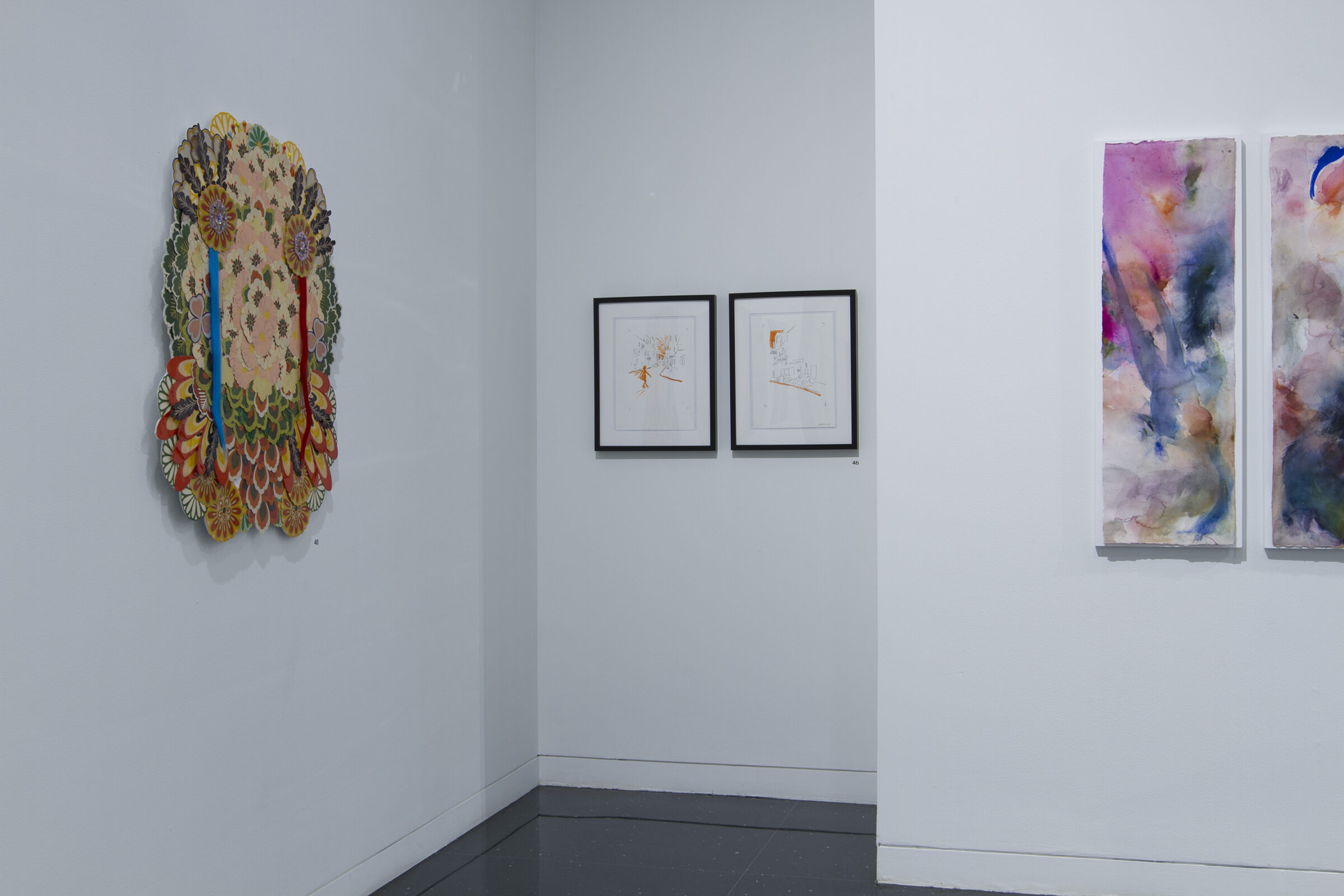 A white walled gallery space in which three artworks hang on the walls. From left to right: an oblong, colorful collage of flowers form a kaleidoscopic pattern, two small, framed drawings in a small nook, and an elongated rectangular watercolor painting of abstract form.