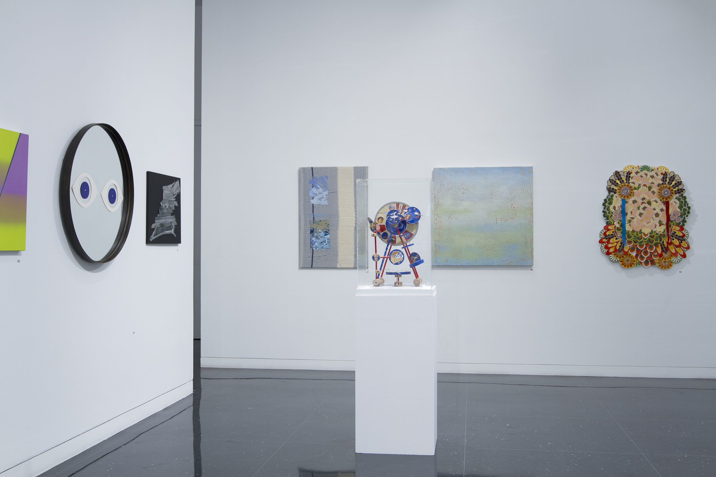 A white walled gallery space in which several paintings and framed artworks are aligned horizontally on the walls and a small, colorful, carnivalesque sculpture stands on a white pedestal