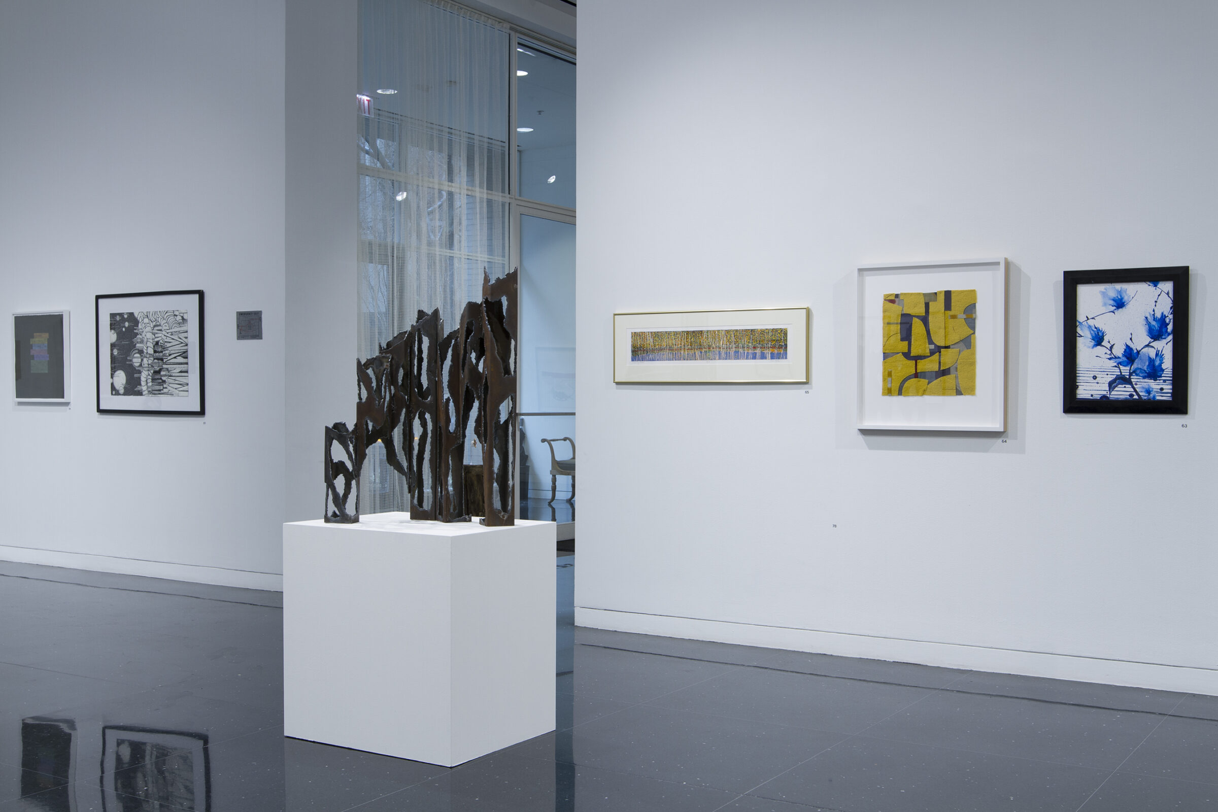 A white walled gallery space in which several paintings and framed artworks are aligned horizontally on the walls and a large abstract metal sculpture resembling an architectural form sits on a cube-shaped pedestal in the middle of the gallery.