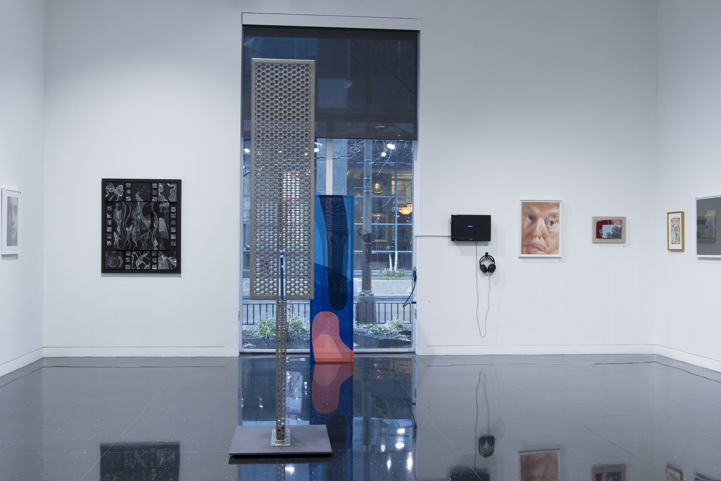 A white-walled gallery with a large window in the center. A colorful abstract fabric sculpture sits in the center of the window, and a large metal sculpture perforated by a grid of holes stands in the gallery closest to the viewer.