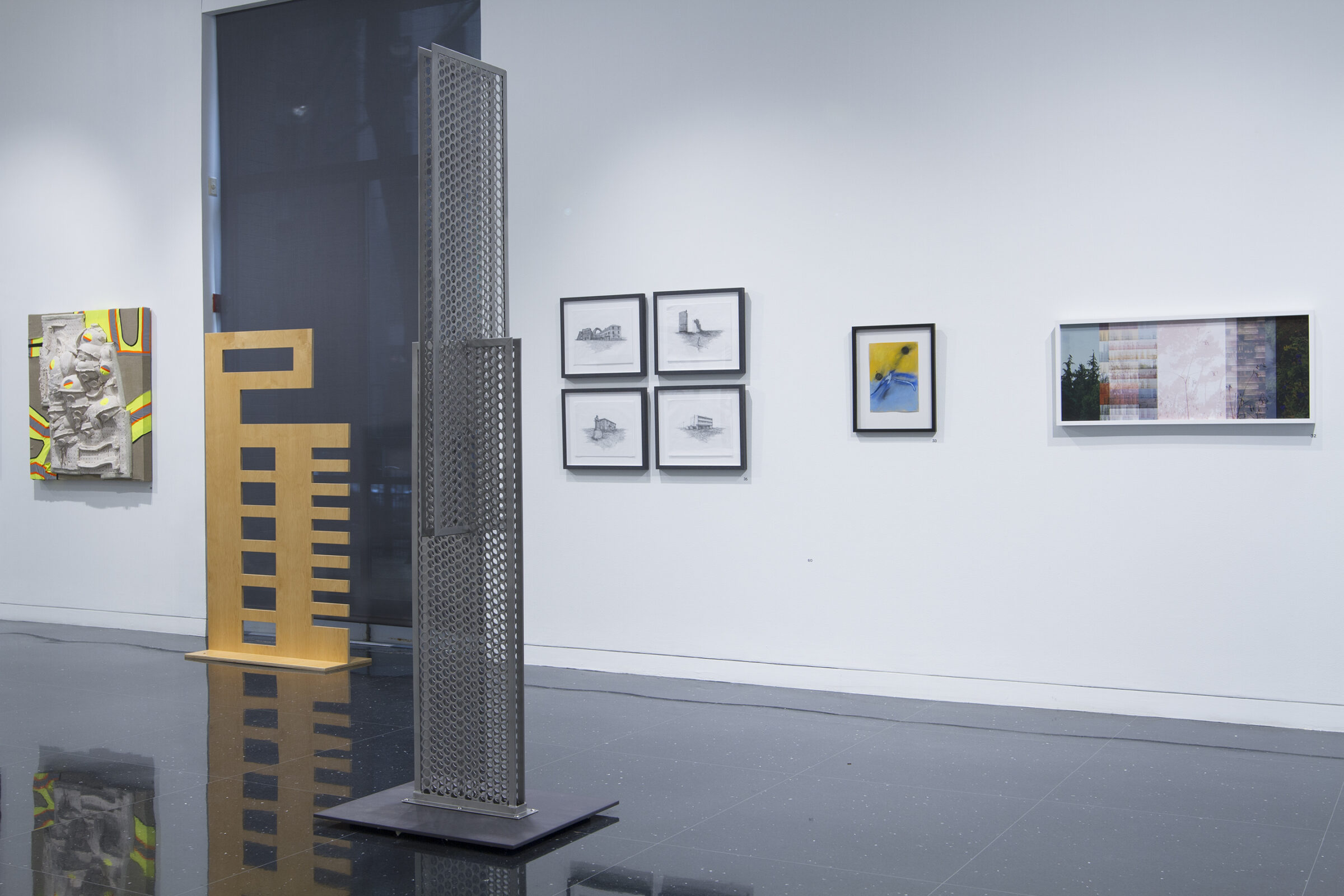 A large metal sculpture perforated by a grid of holes stands in the white gallery closest to the viewer. Behind it are four small framed images arranged in a grid, and next to those, framed by a large window is a wood sculpture with square holes and rectangular notches cut out of it.