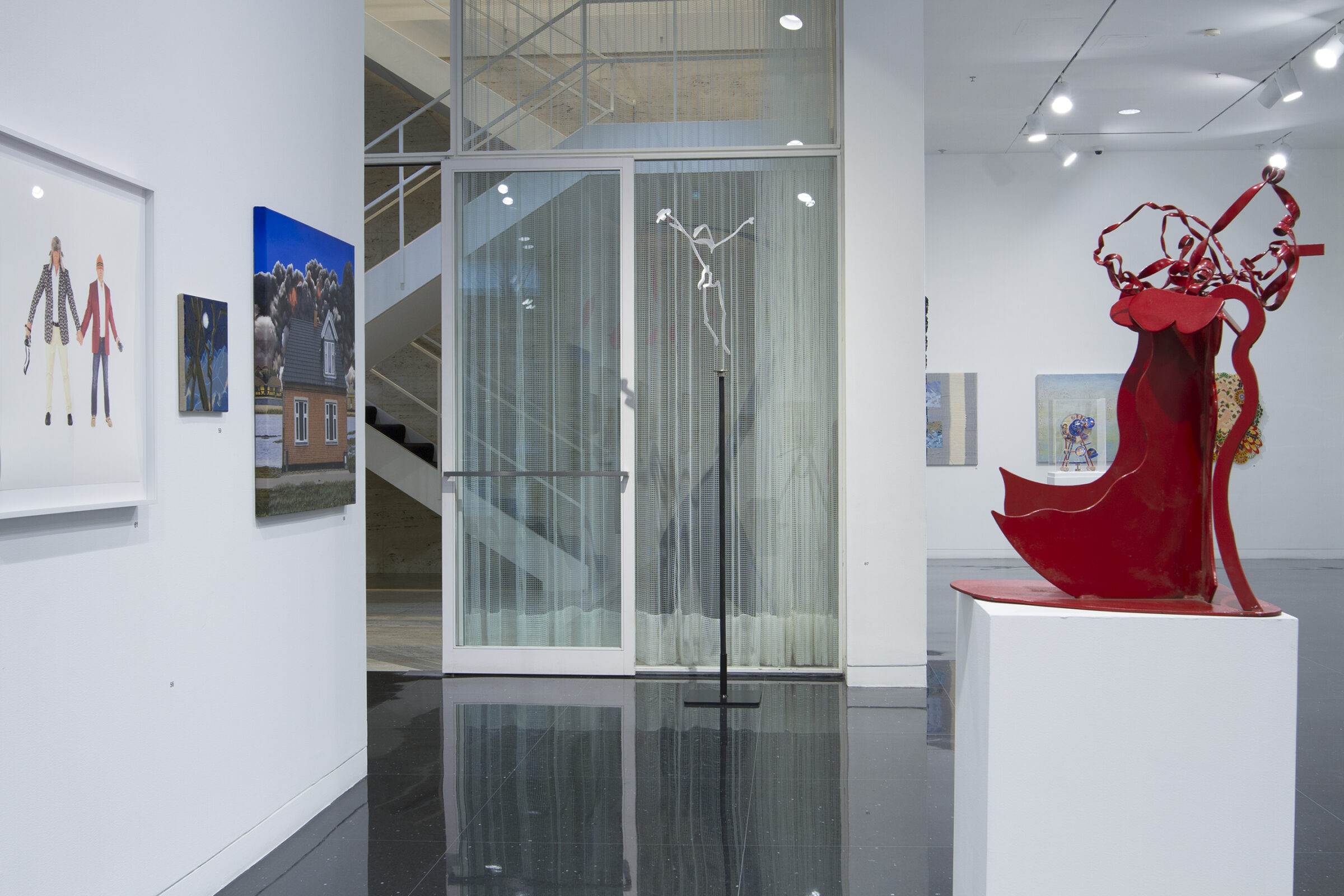 A white gallery space in which a red steel sculpture of an abstract human form sits on a white pedestal across from a framed photograph and a painting of a house. In the distance, a white, wire sculpture is installed to appear balancing on a tall, thin pedestal. The Mies van der Rohe stairwell is also visible.