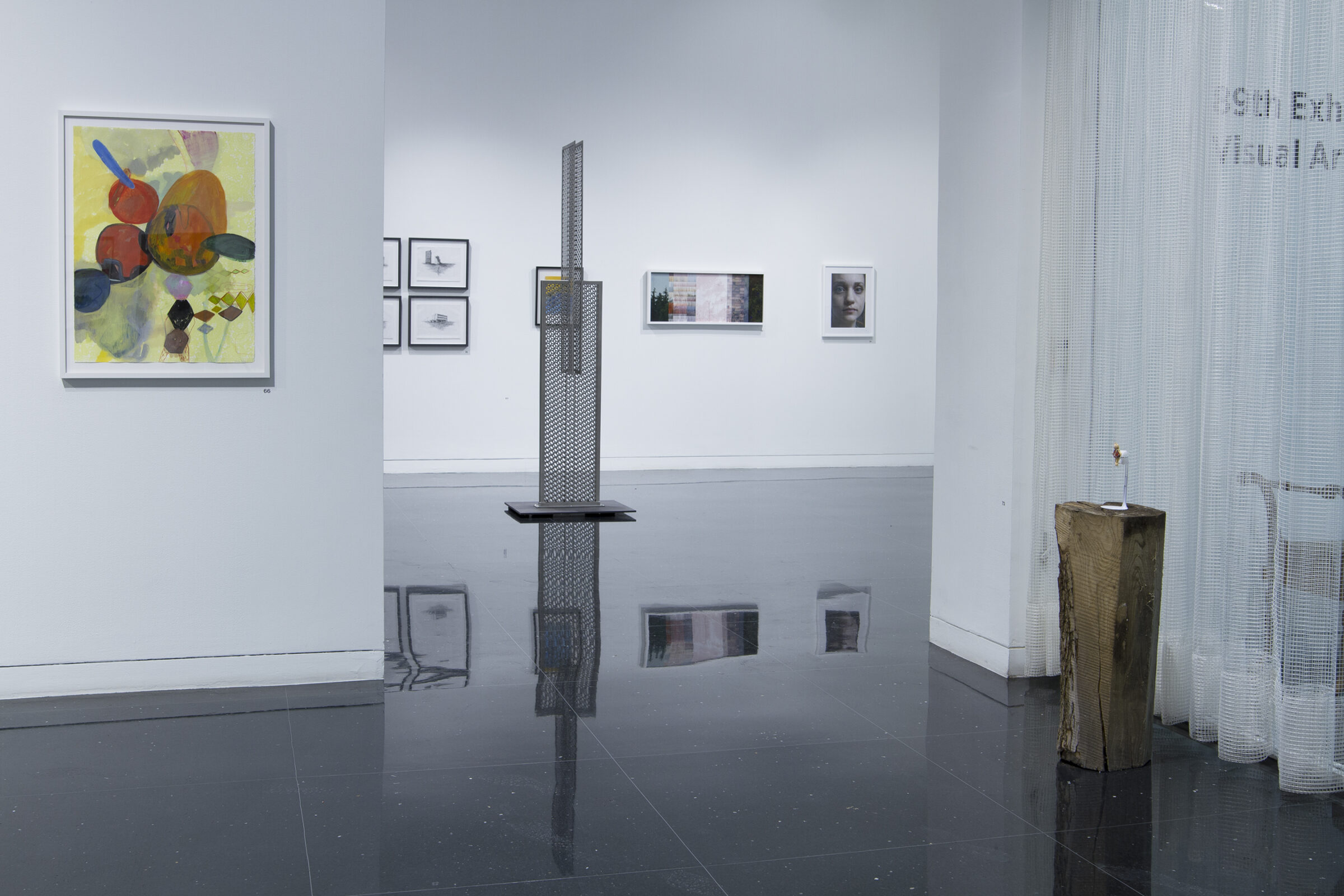 A white gallery space where a very small sculpture stands on a large wooden log to the viewer's right, across from which hangs a colorful, framed abstract painting on a freestanding wall. A large metal sculpture perforated by a grid of holes stands in the distant gallery among other framed wall works.