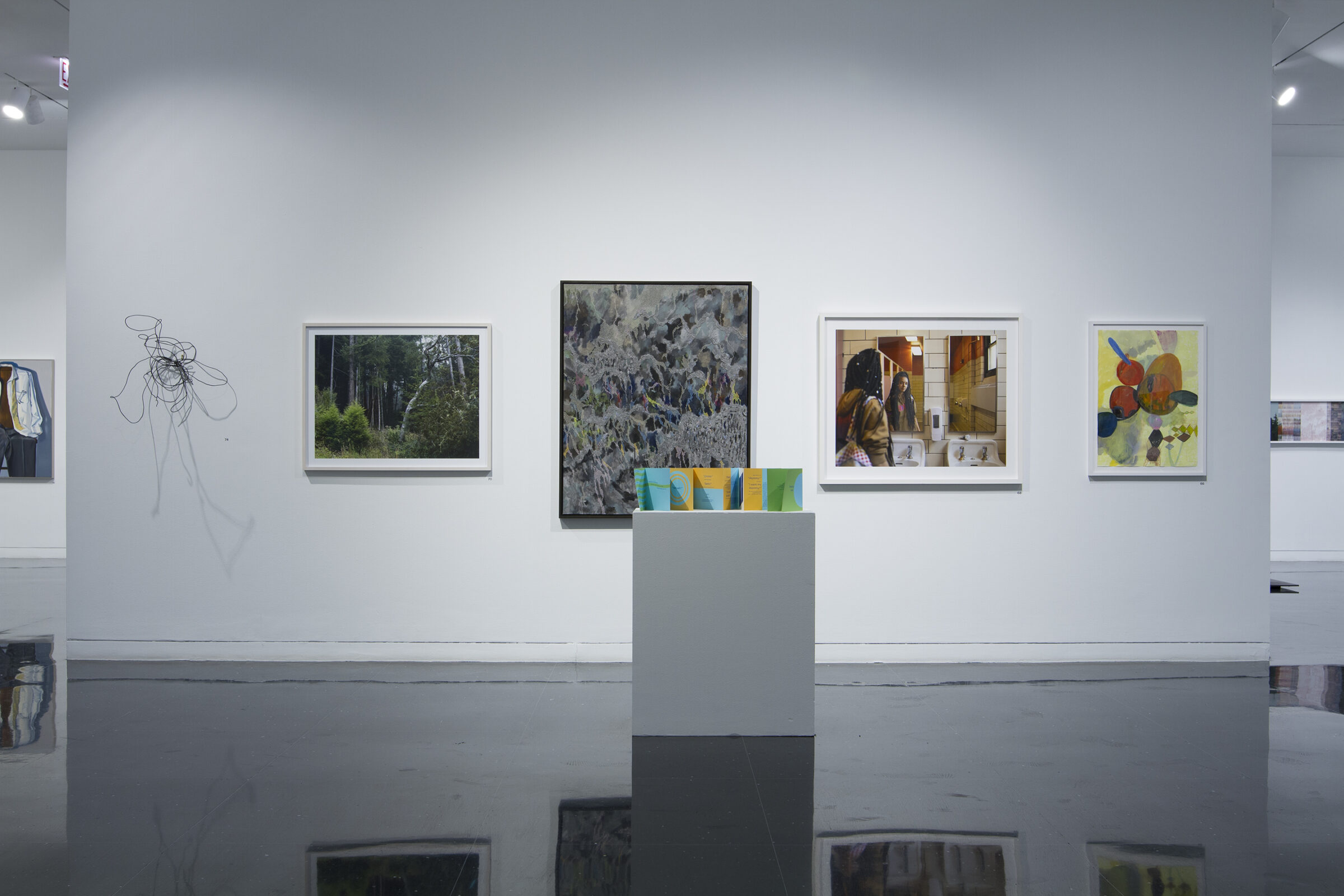 A white, freestanding gallery wall holds four framed artworks arranged horizontally and one abstract wire sculpture resembling a tangle or nest on the far left side. In front of the wall stands a paper, accordion-folded pamphlet printed on green, blue, and golden yellow paper.