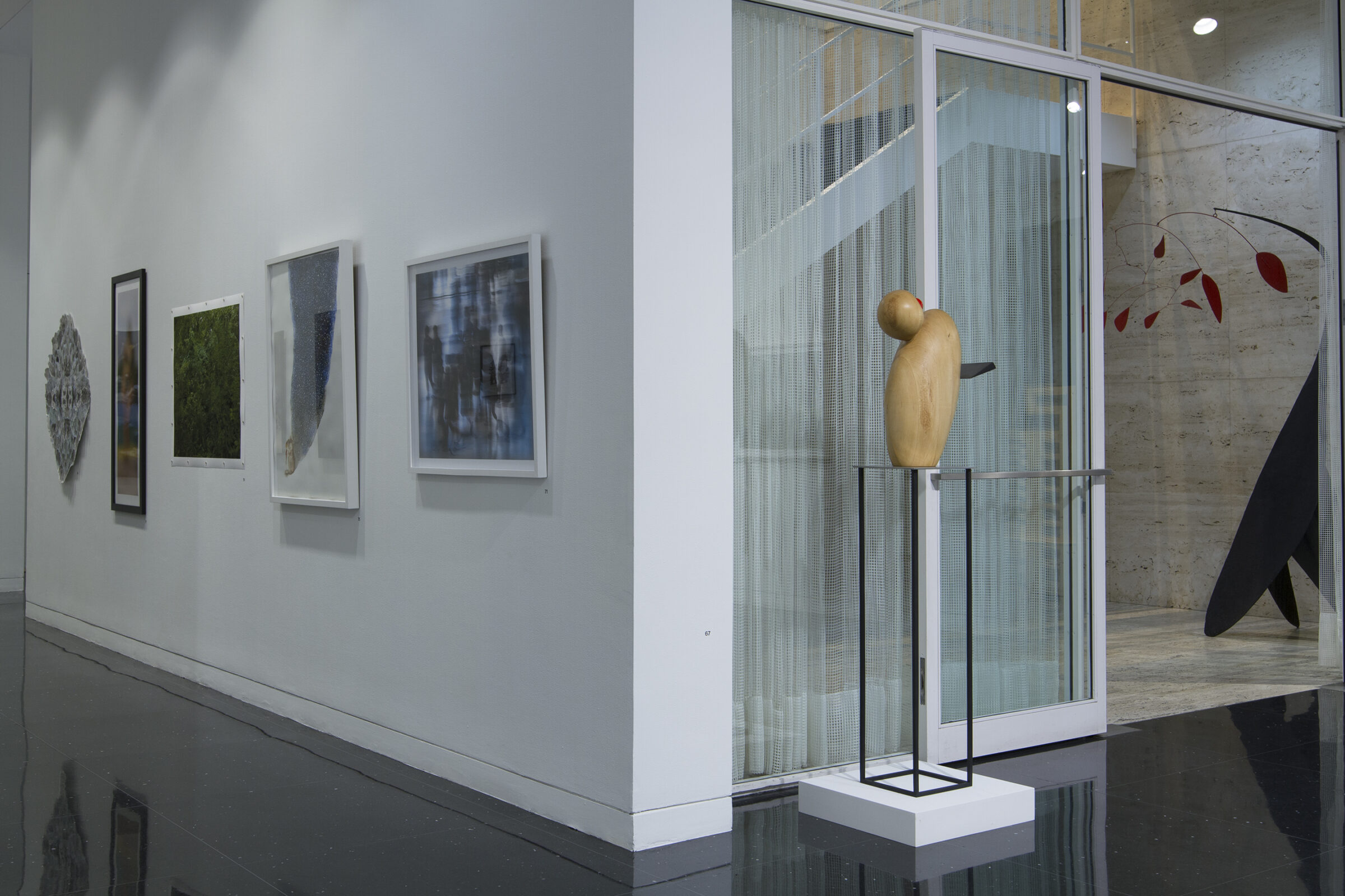 In a white gallery, a wooden sculpture of a duck or birdlike creature stands on a black wire pedestal in front of the Mies van der Rohe stairwell. To the left of the sculpture and around the corner, five artworks are arranged in a horizontal line down a hallway.