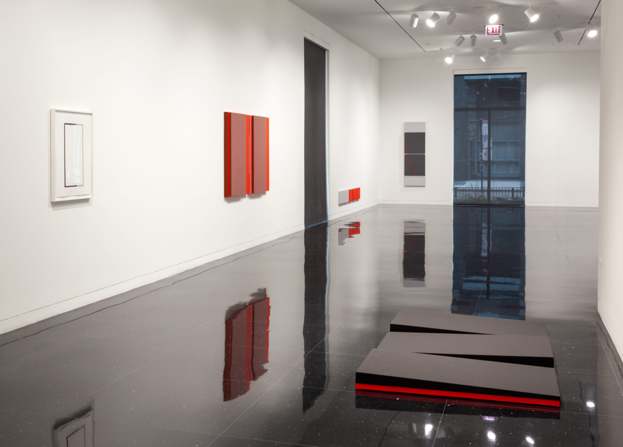 Installation view of a gallery with abstract red and gray artworks on the white walls. On the floor in the foreground is a series of 3 black sculptures with bright red bases that resemble ramps.