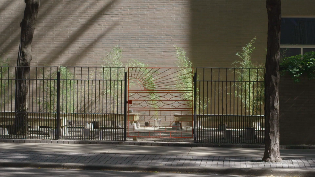 A black garden fence with an ornate red gate stands in front of a small brick-walled garden.