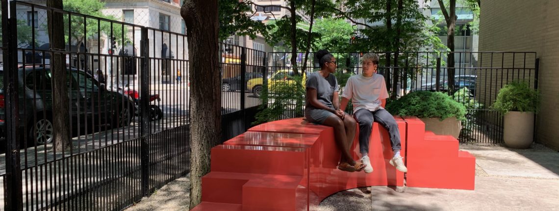 Two people sit on large red box-like furniture in an outdoor patio.