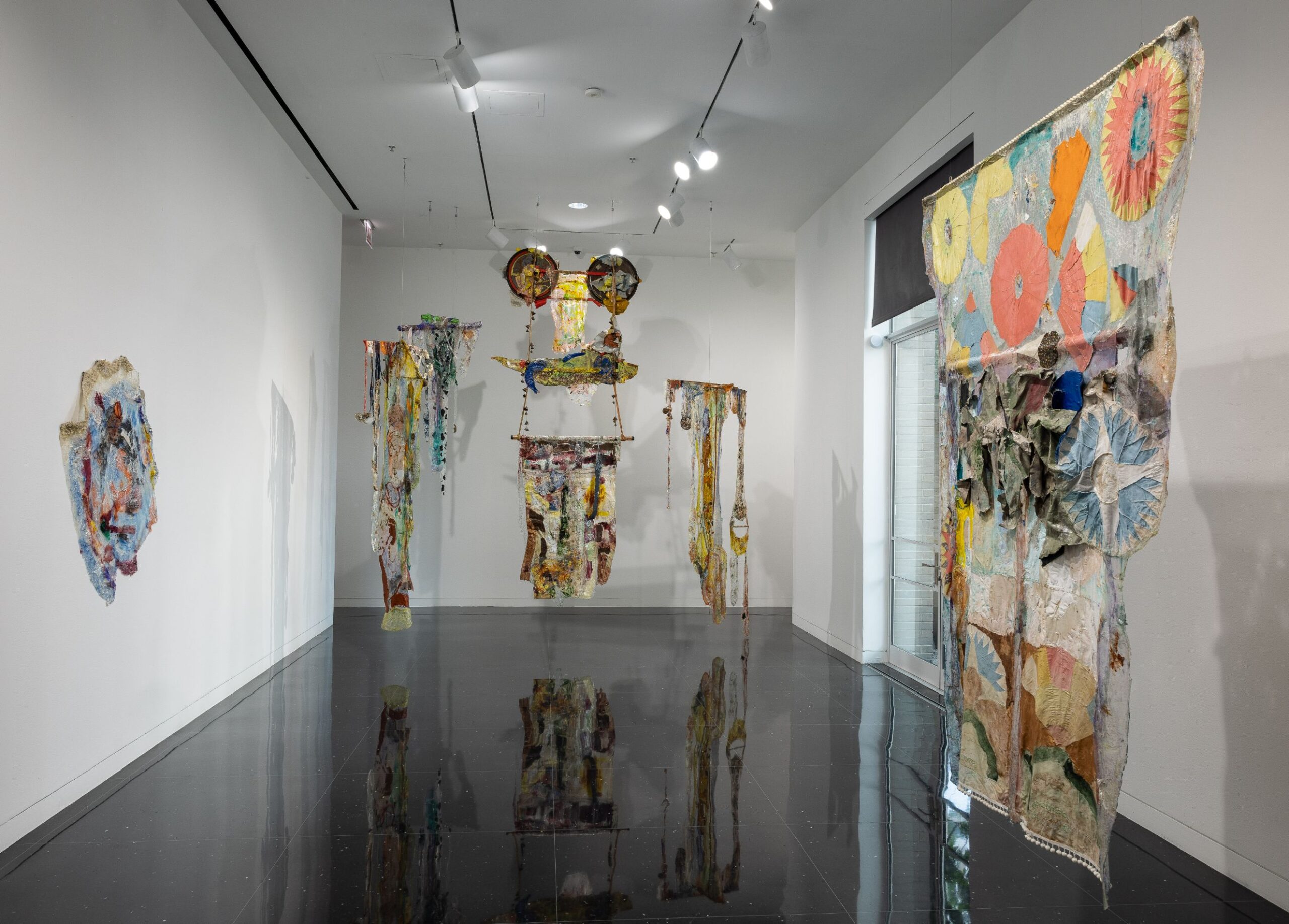 A large white gallery in which six colorful, mixed media artworks hang from the ceiling.