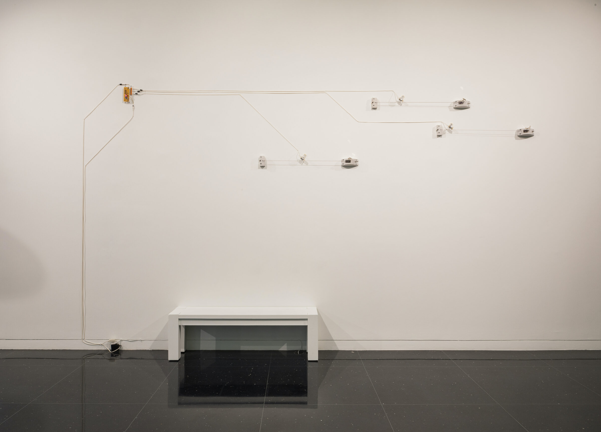 A series of nodes and wires installed on gallery walls. Underneath the installation sits a white bench.