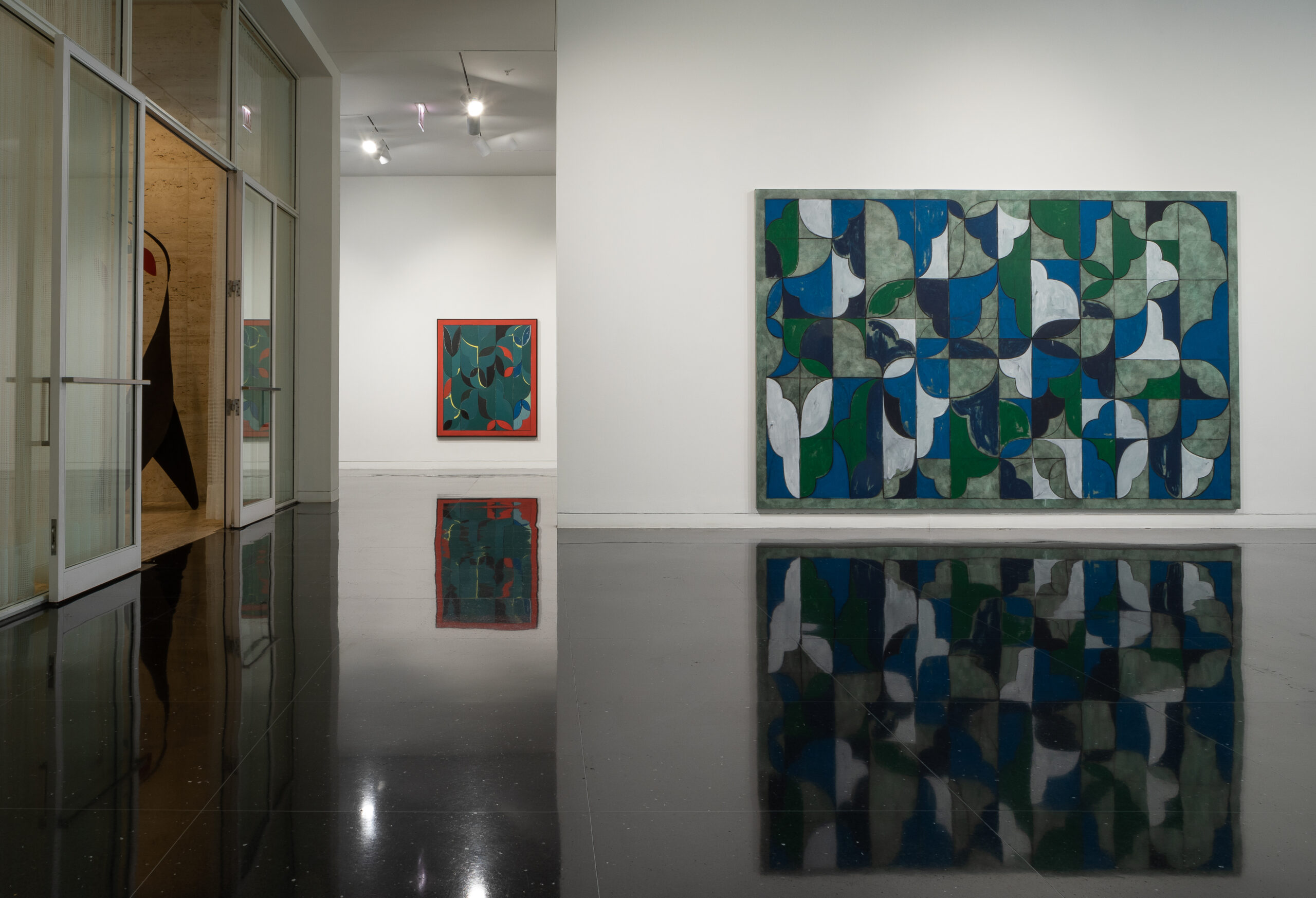 A large, nearly wall-sized abstract painting with blue, white, gray, and green interlocking shapes displayed on a white gallery wall. To the left of this painting in the distance, another abstract painting is visible on a far wall.