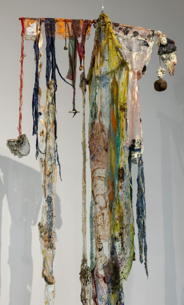 A colorful mobile made of fabric scraps and a transparent material hangs in front of a white wall.