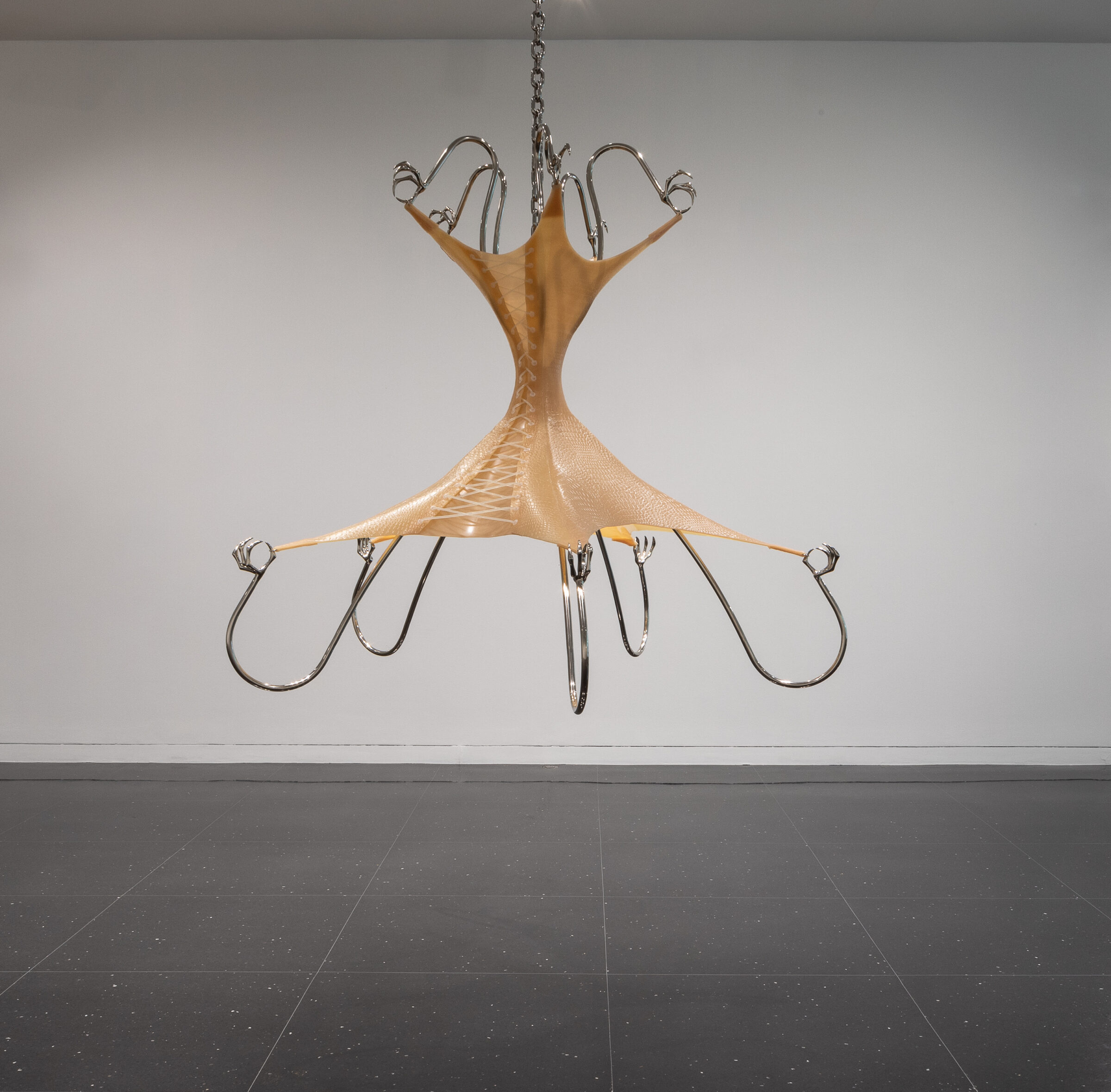 A steel and flesh-toned latex sculpture resembling a mix between a corset and a chandelier is suspended in a white gallery space.