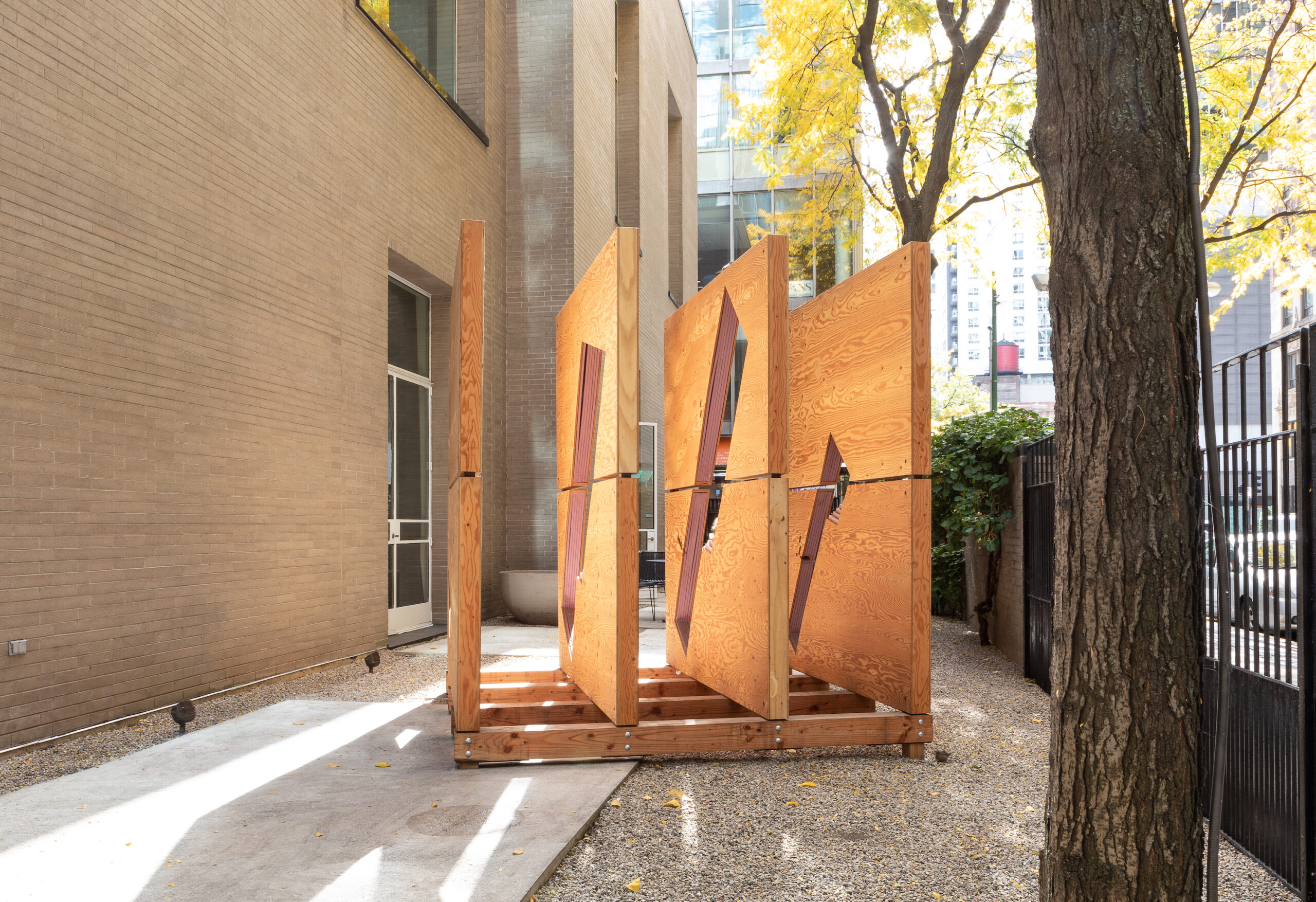 Four wood slabs resembling walls of a crate viewed from the side installed on a wood pallet on the concrete patio of a garden. To their left, the brick exterior of The Arts Club of Chicago is visible.