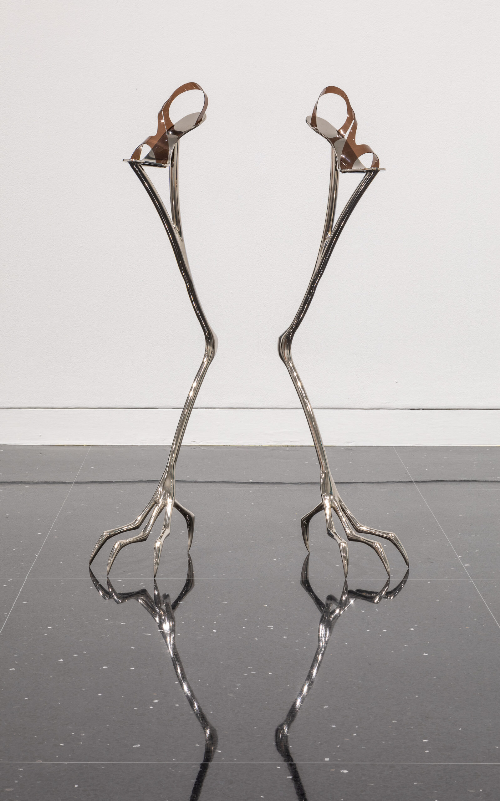 A pair of steel stilts with sandal-like foot straps and base supports resembling bird feet stands in a white gallery.