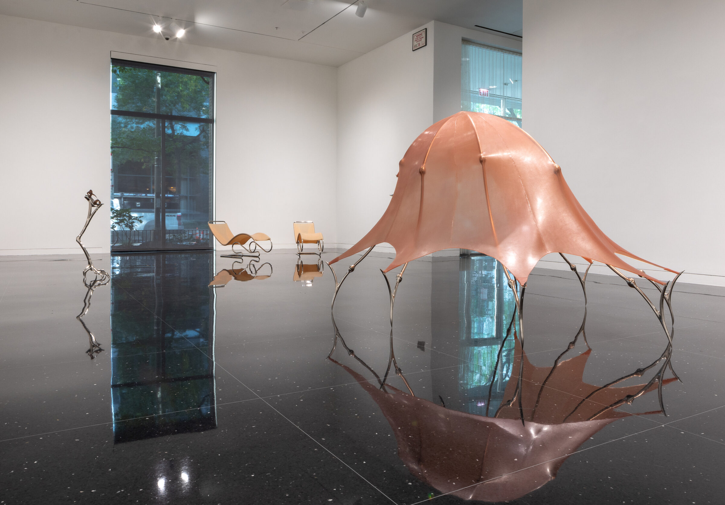 A large sculpture made of steel and latex and resembling an umbrella shape stands in a white gallery. A pair of steel stilts and two lounge chairs, also in latex and steel, are visible in the background.