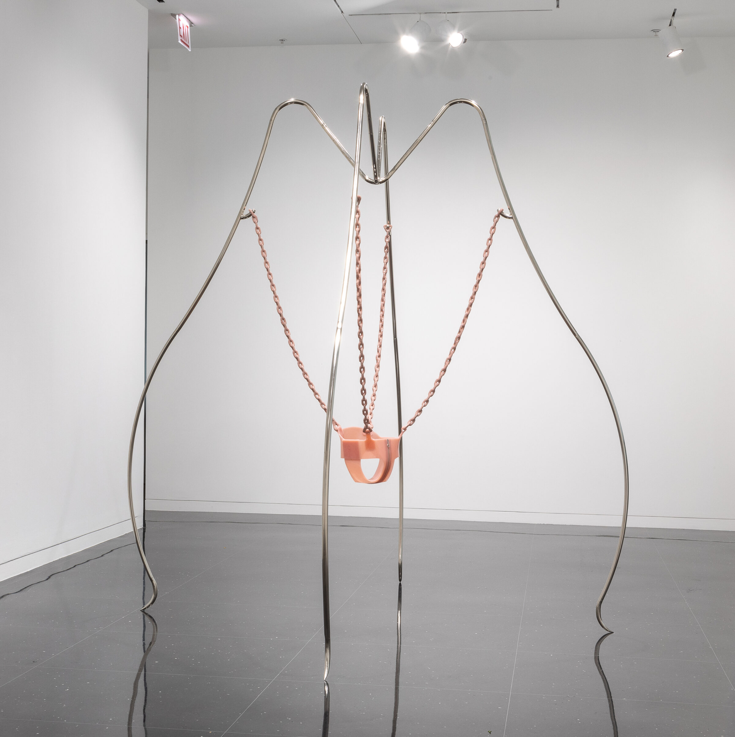 A white gallery with a steel and latex sculpture installed in the middle. The latex component is shaped into a toddler's swing and is suspended by four long steel supports bent like insect legs.
