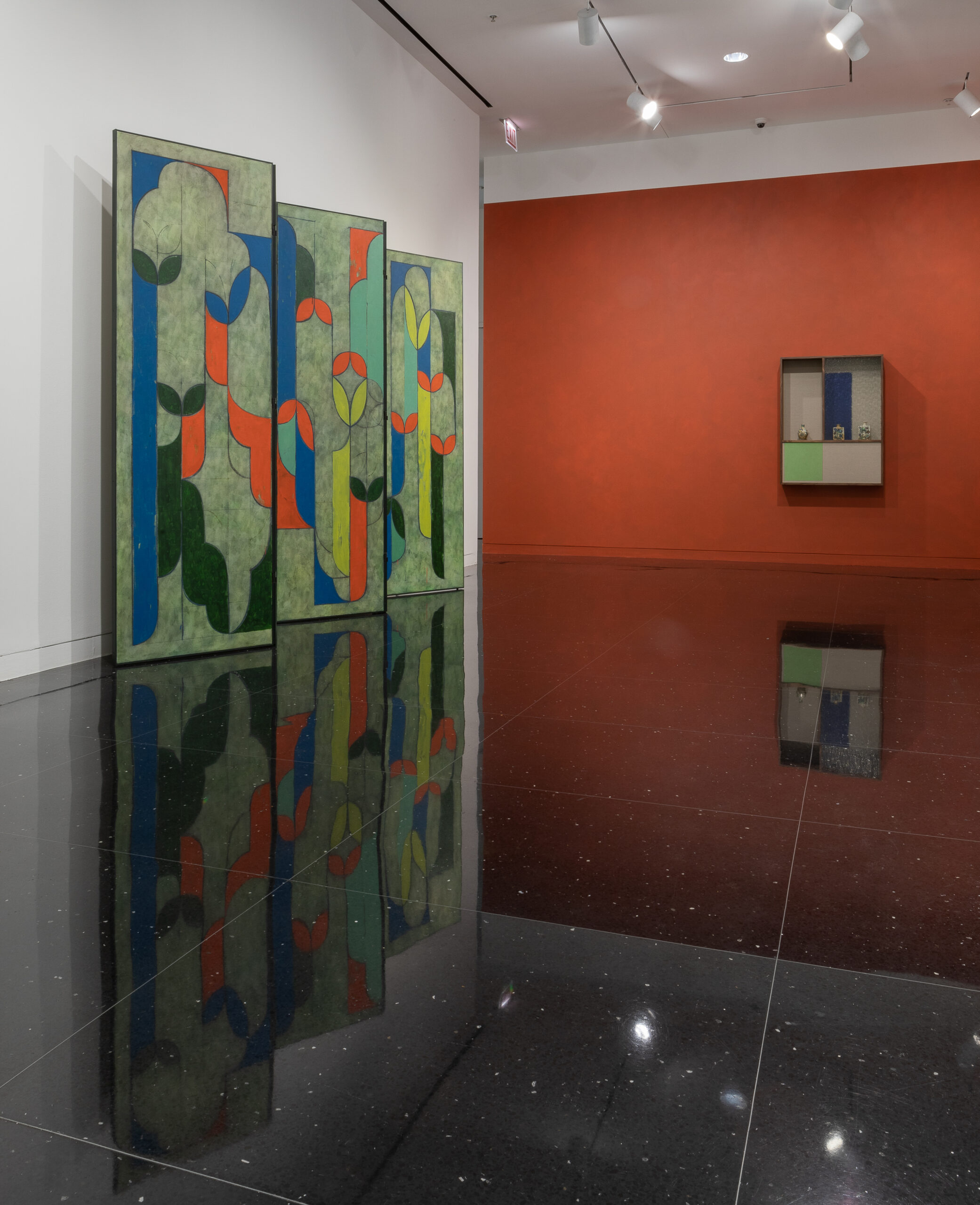 A gallery space with one wall painted a deep orange. To the left of the wall stands a 5-panel screen painted in a green and orange abstract pattern.