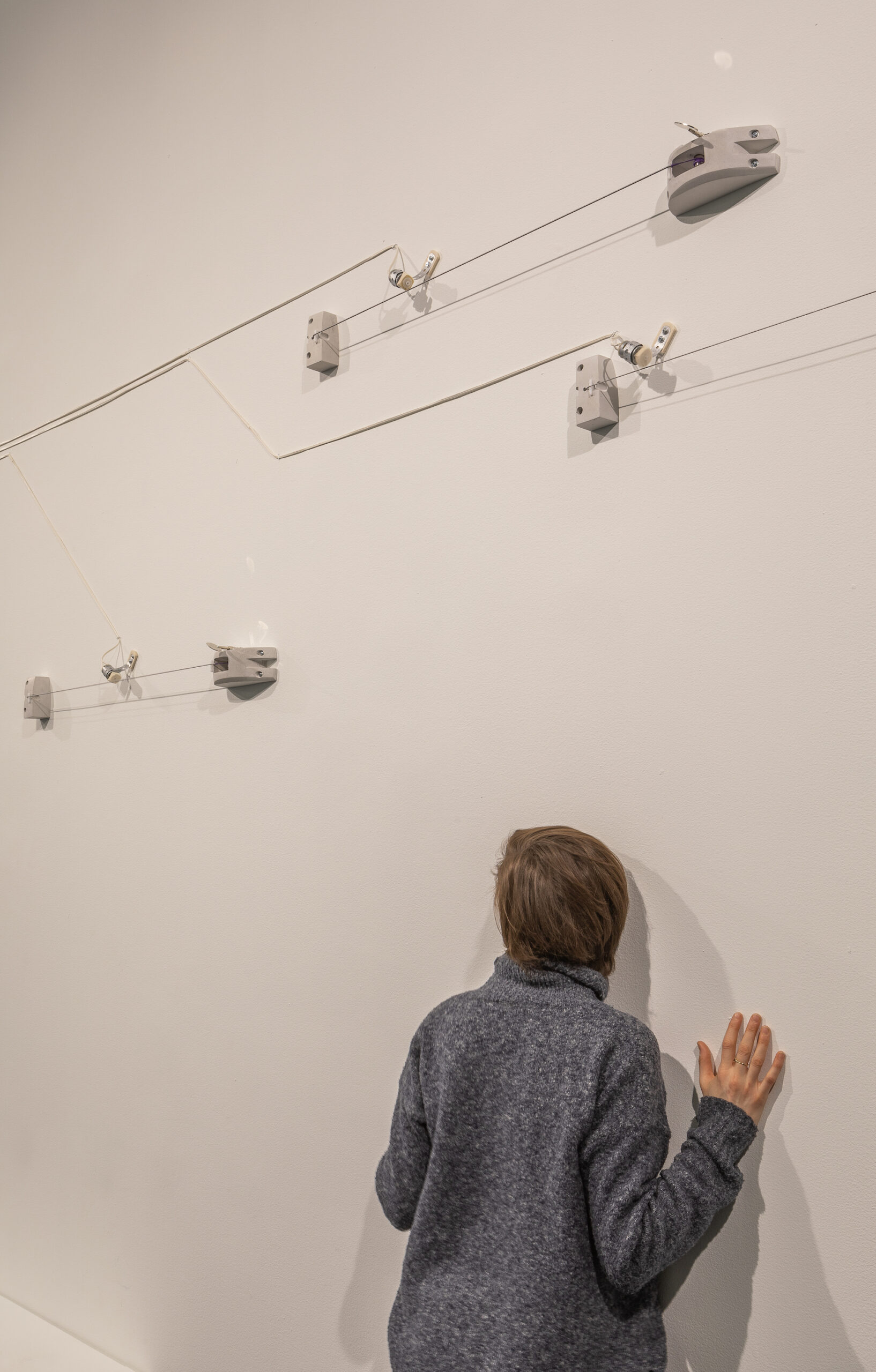 A series of nodes and wires installed on gallery walls. A woman stands underneath the installation with her face and palms against the wall.