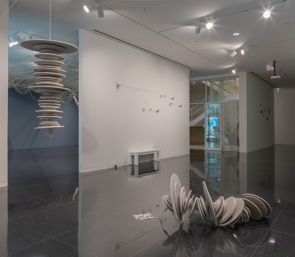 To the left, a series of off white disks of various sizes suspended from the ceiling with a white rope. To the right, a series of off white disks of various sizes strung together with a white rope and placed on the gallery floor.