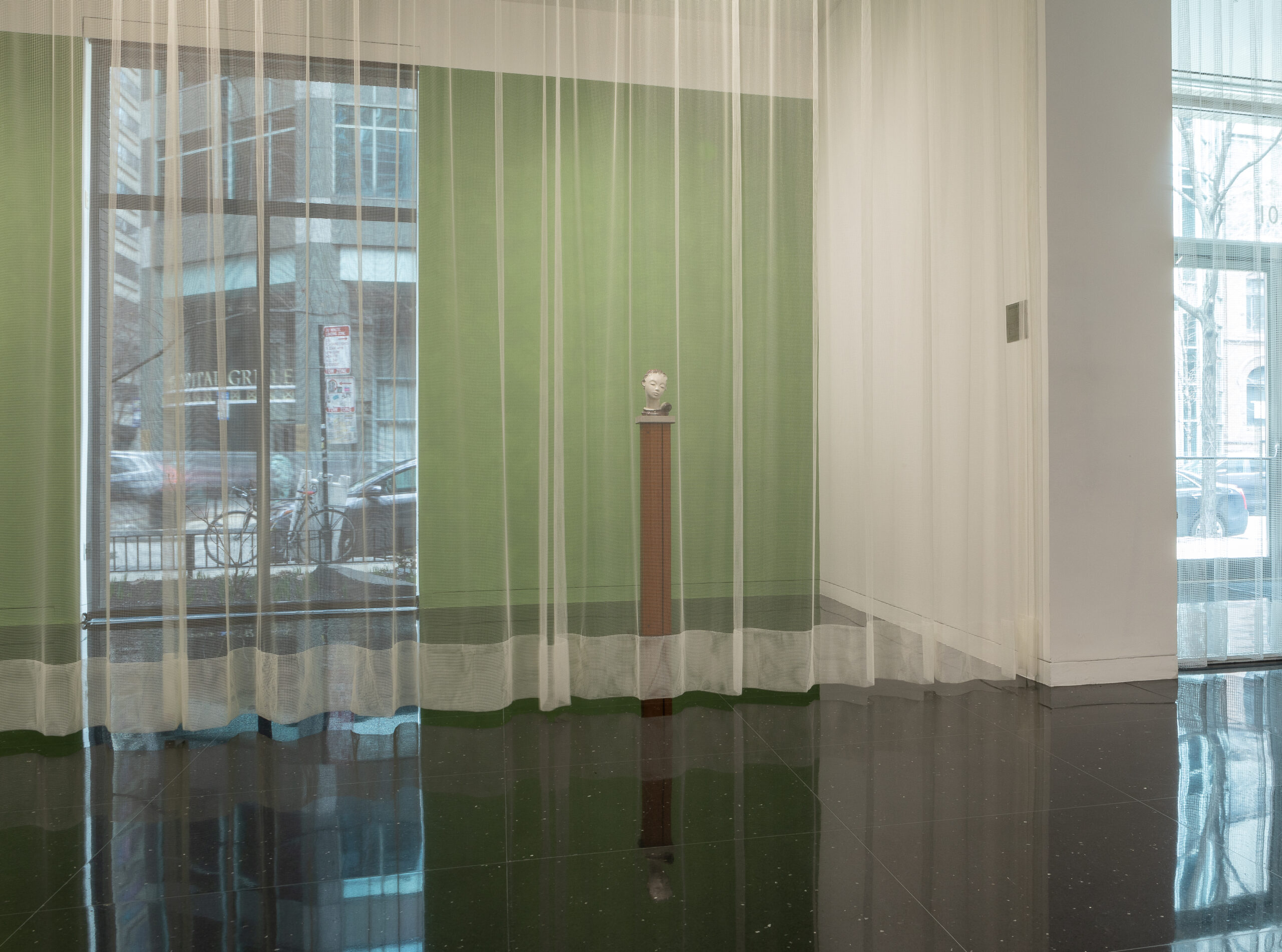 A sculpture of a head sits on a brown pedestal in front of a gallery wall painted pea green. In front of the statue is a transparent white mesh curtain that spans the length of the gallery.