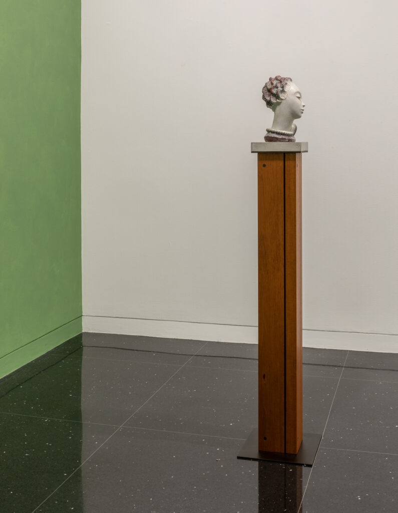 A sculpture of a human head on a brown pedestal. Behind the sculpture, one gallery wall is painted pea green.