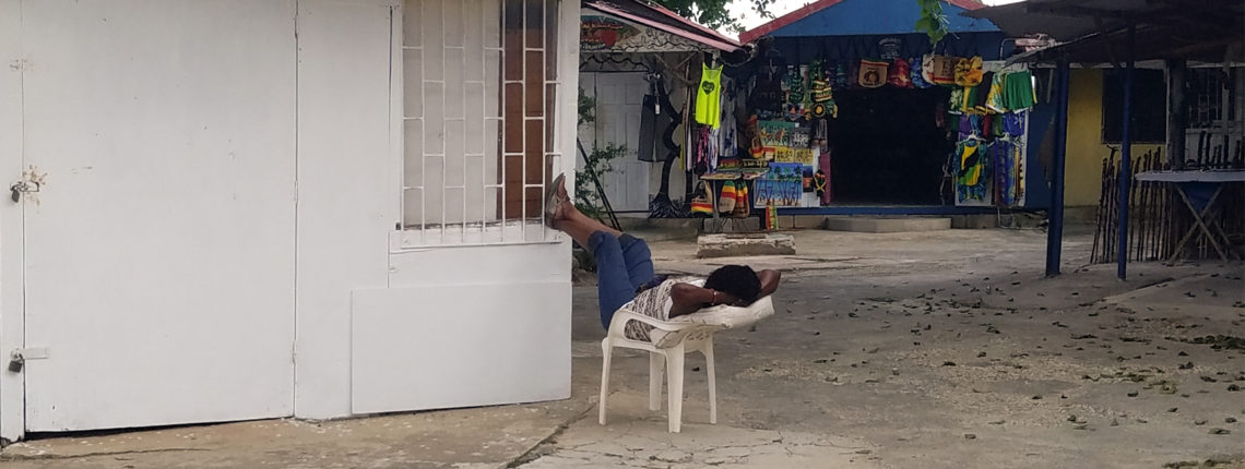 A Black person lounges outdoors in a plastic chair, their feet up on a window ledge.