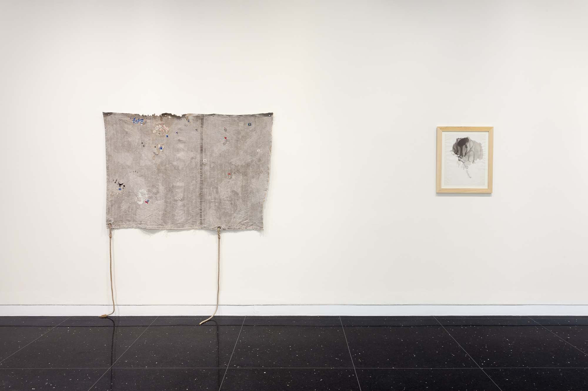 A pale gray, somewhat ragged looking textile hangs on a white gallery wall next to a small, square image in a white frame.