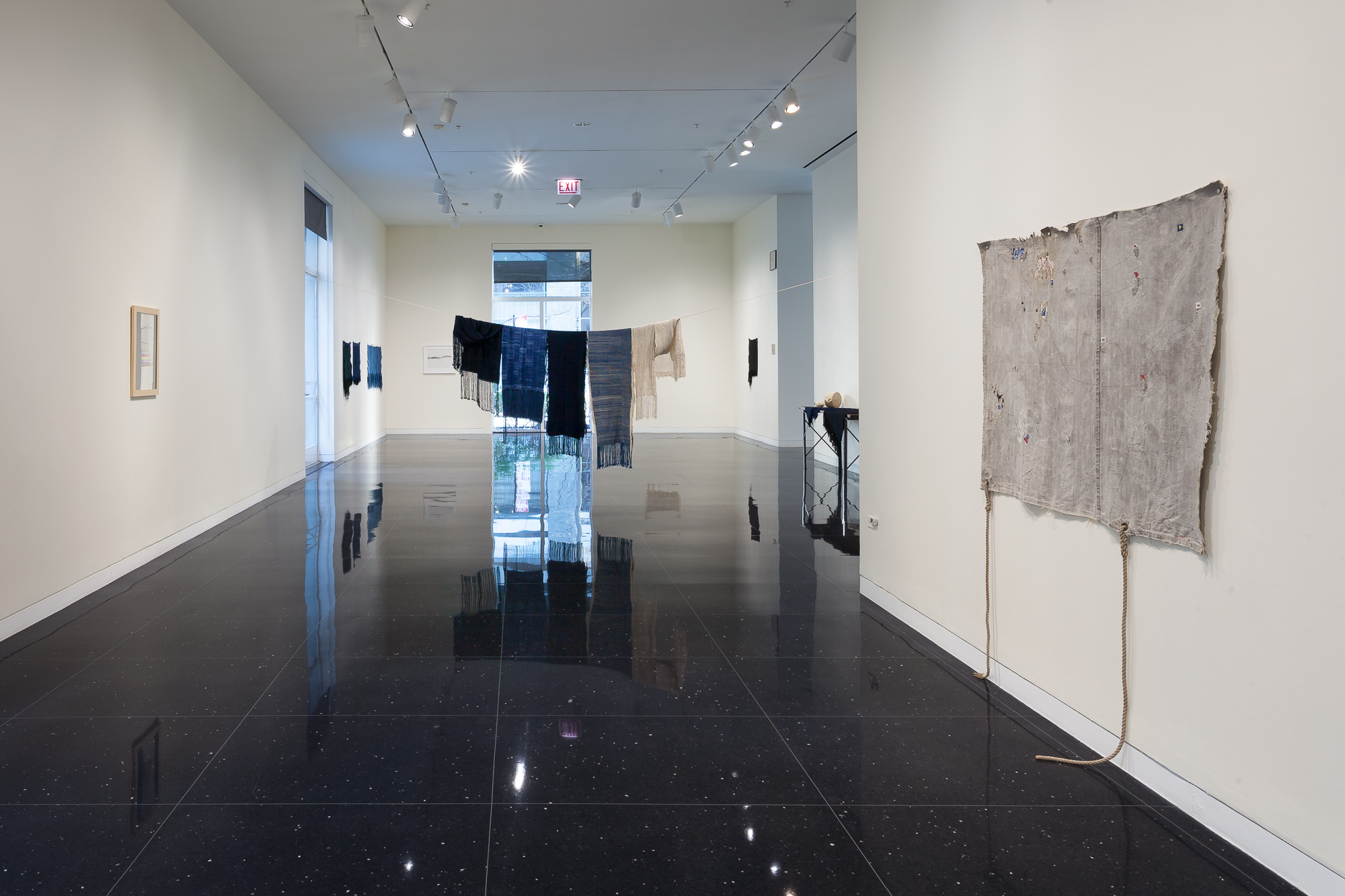 Installation view of a white walled gallery with several installation of textiles. A pale gray, slightly fringed textile hangs on the right wall. In the middle of the gallery hang several indigo dyed cloths suspended on a clothesline.
