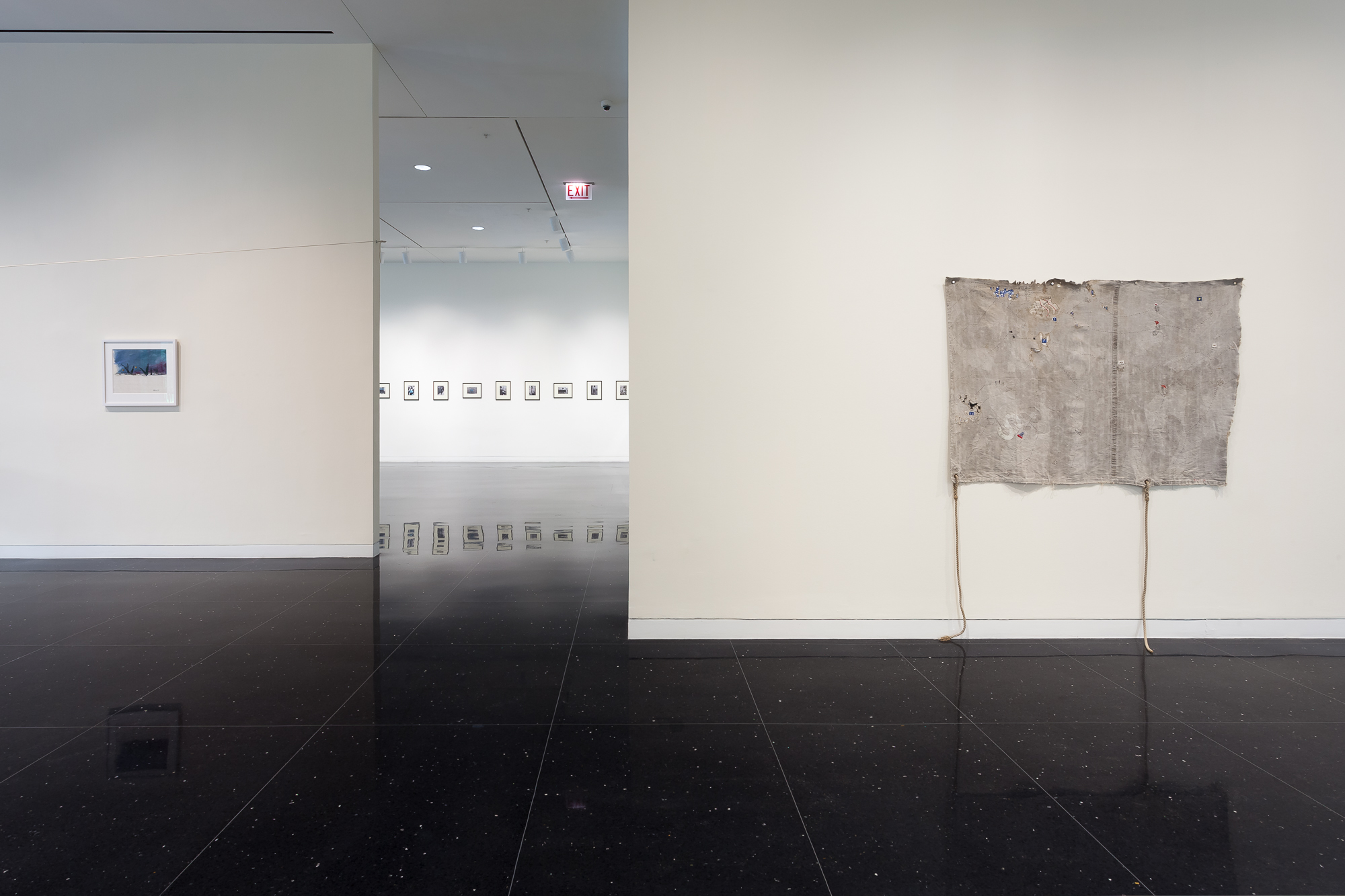 A freestanding white gallery wall with a pale gray, somewhat ragged looking textile hanging from the wall. On a far wall, a line of small, square, framed images hang in a horizontal line on the wall.