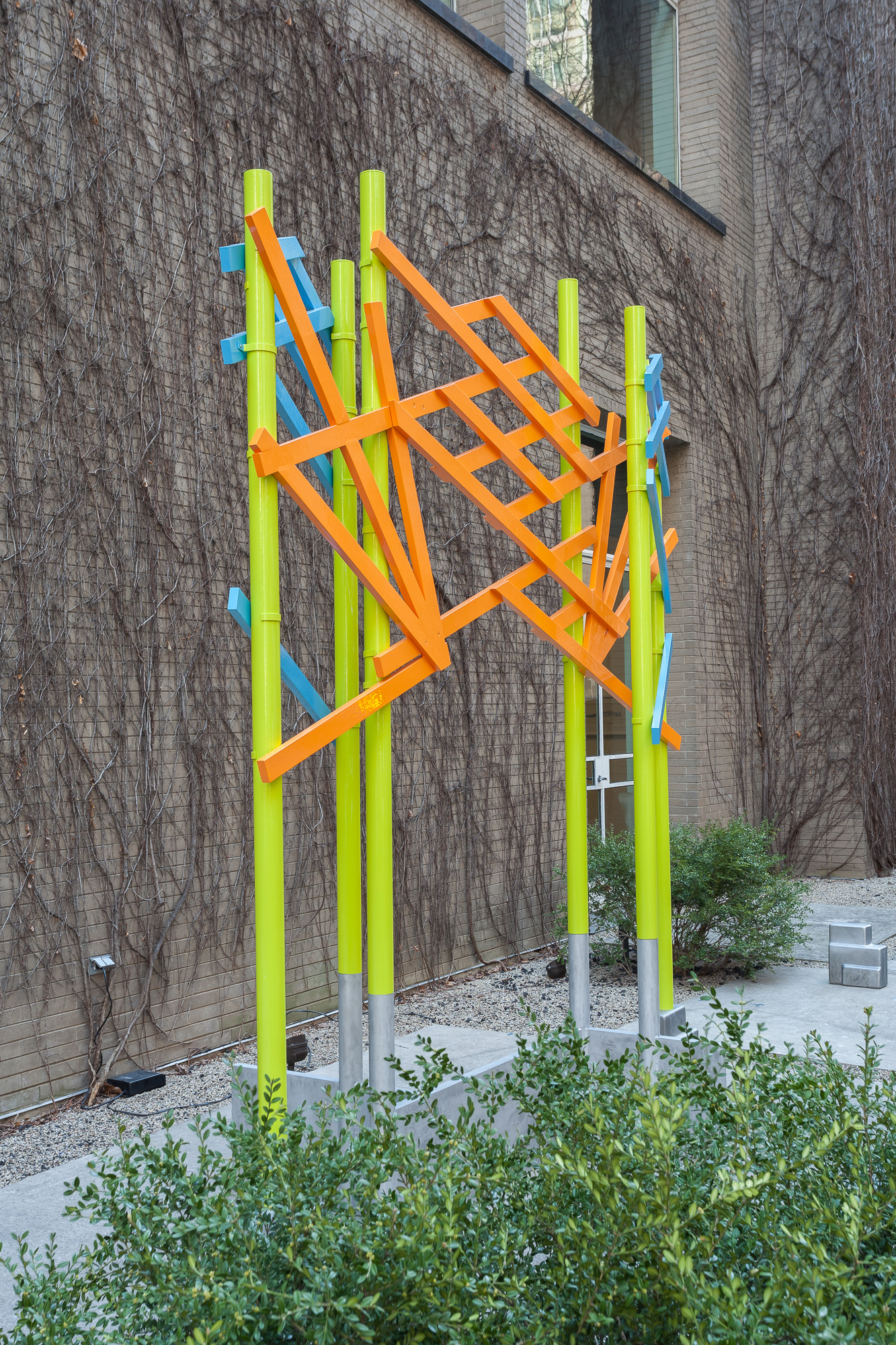 A large shrub in front of six bright green poles attached to the ground with cast metal bases. The green poles are interconnected through bright orange and blue latticework, forming an arch or gate.