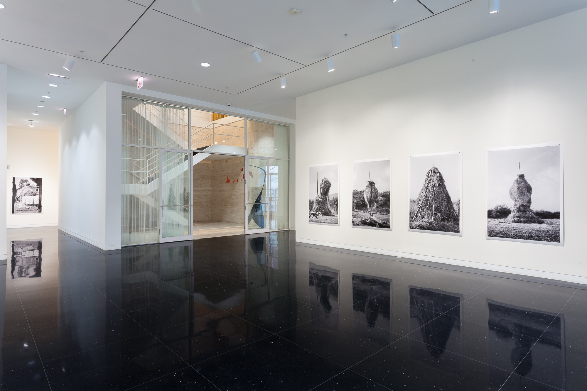 A freestanding white gallery wall with four life-size black and white photos of haystacks shaped into organic, animal-like or humanoid figures. To the left of the wall, the white Mies van der Rohe-designed stairwell is visible.