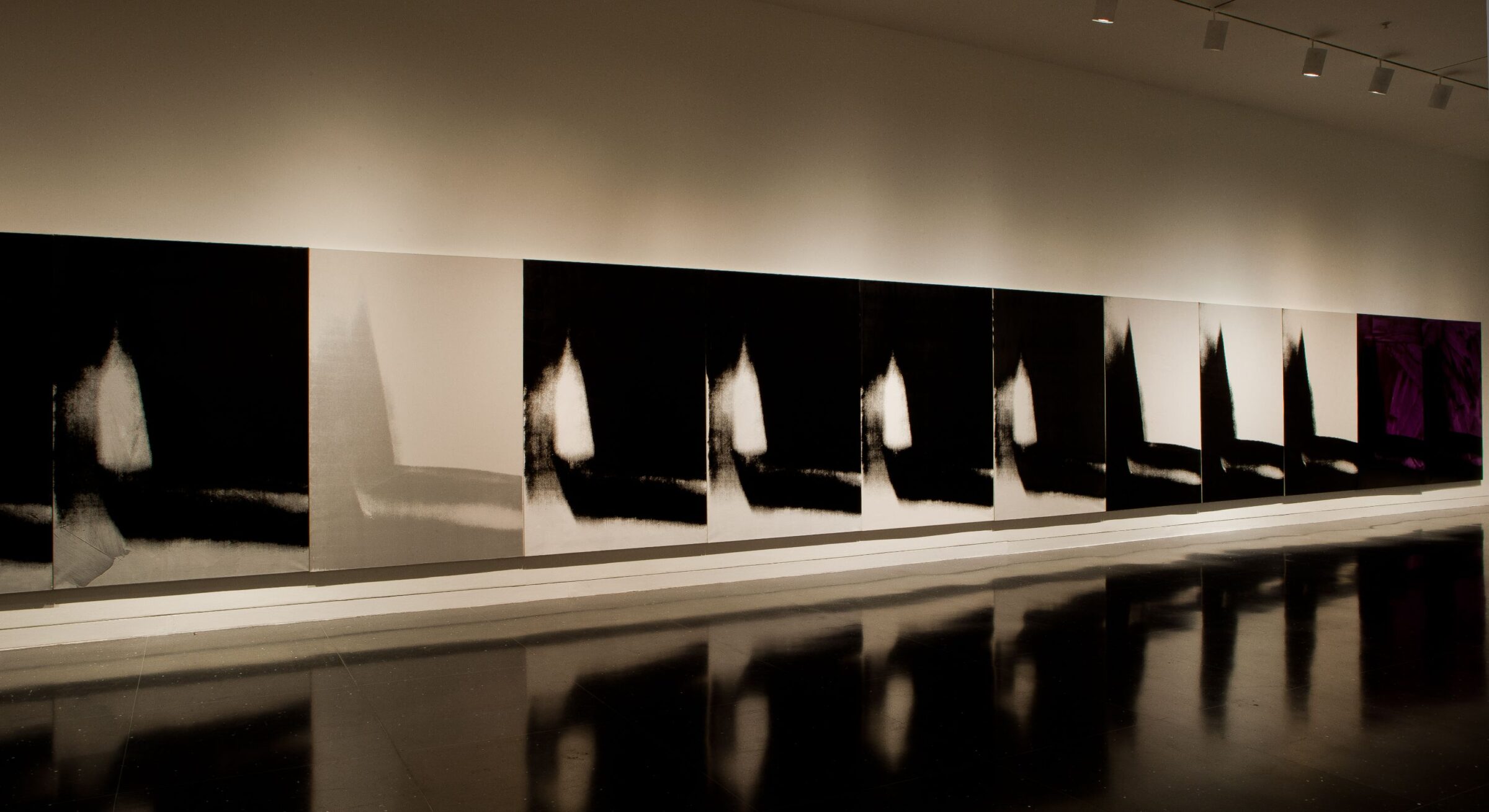 Several mostly black paintings of an identical abstract image in black and white span the length of a white gallery wall.