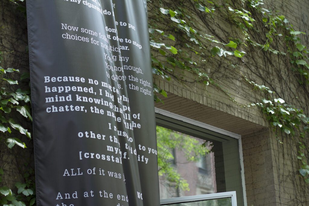 A black banner with text in various shades of gray hangs on a brick wall covered in ivy.