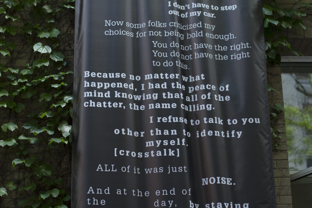 Close up of a black banner printed with text in various shades of gray hanging on a brick wall covered in ivy.