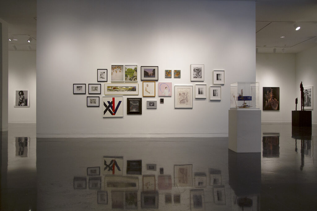 A series of artworks are hung gallery-style on a white wall. More artworks are visible in the next room back.