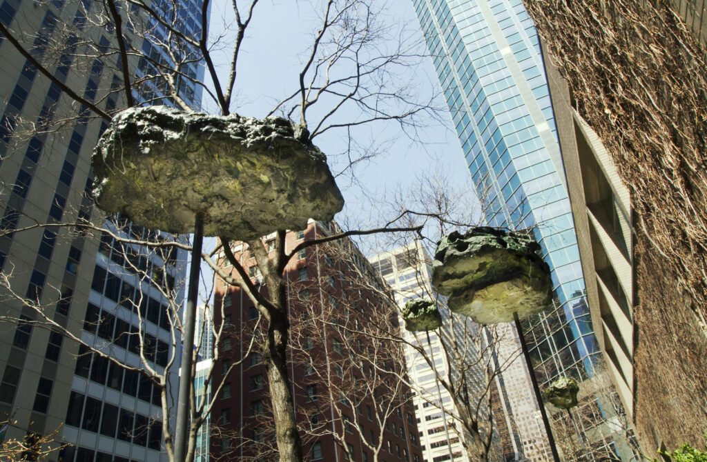 Two large and two small asteroid-like objects with a green tint to them rest atop metal posts in an area filled with skyscrapers.