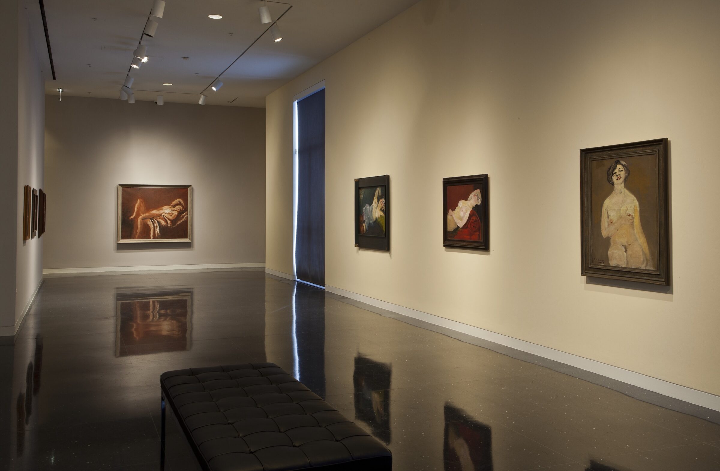 Four framed nude paintings of varying size hang in a beige gallery. The painting furthest from the viewer depicts a reclining nude figure and hangs centered on the wall. A cushioned black bench sits in the gallery closest to the viewer.