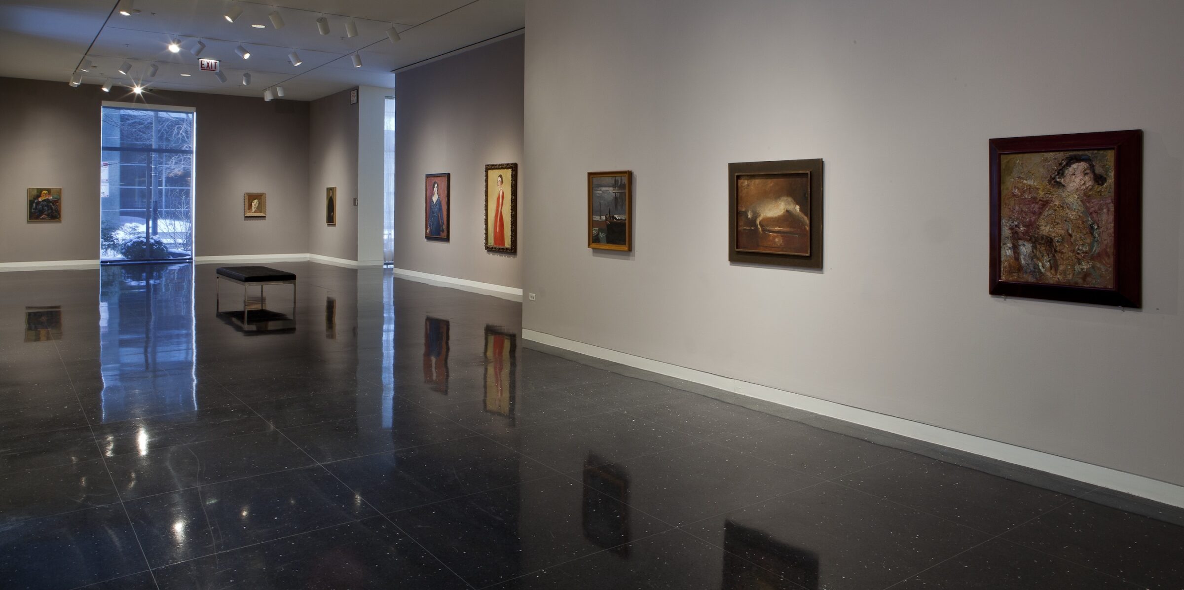Wide view of a beige gallery in which eight framed paintings of various subjects can be seen hanging on its walls. The majority of the paintings depict female figures. The bust-length portrait furthest to the right and closest to the viewer depicts a figure's bust abstracted by thick strokes of paint.