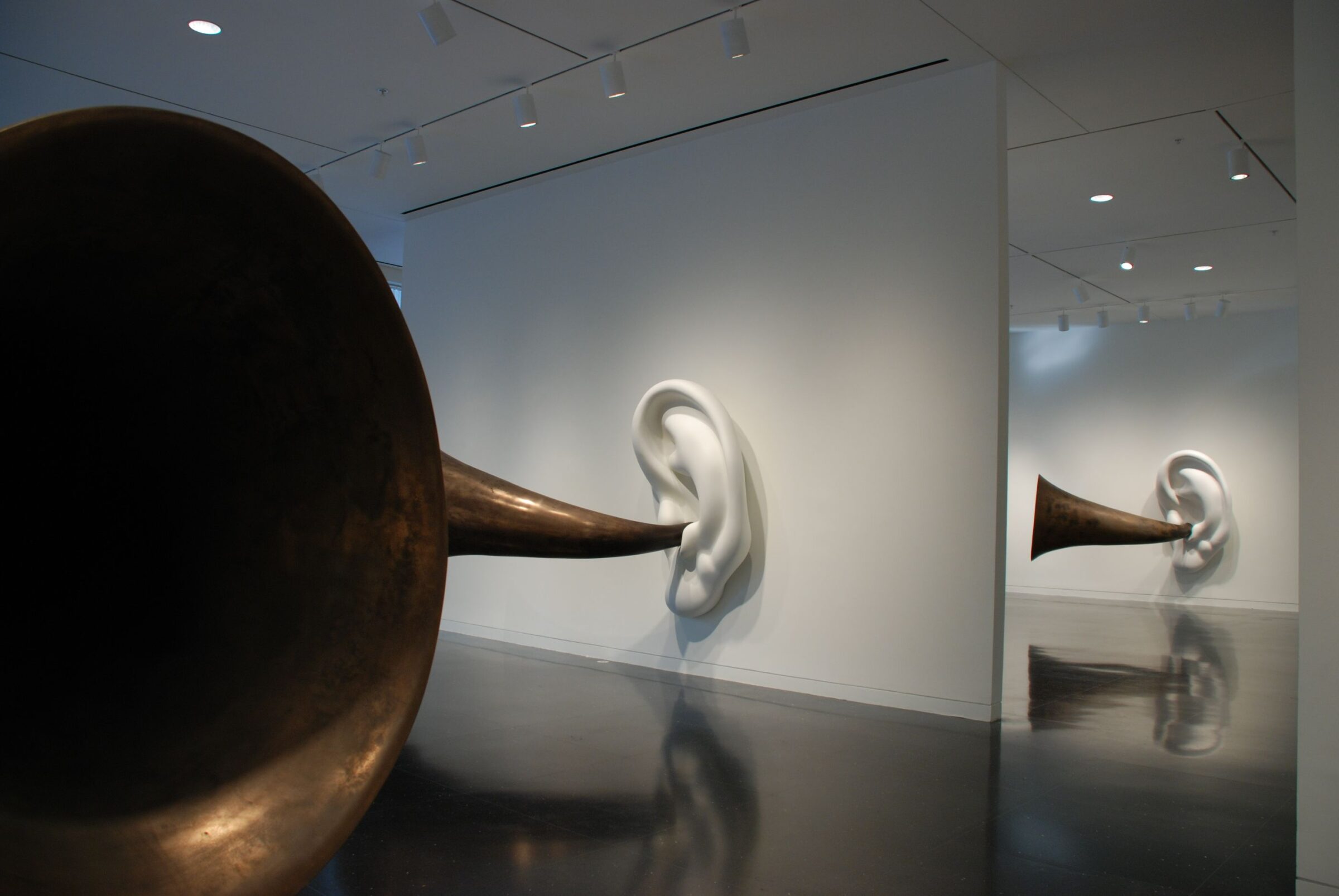 A freestanding white gallery wall holds a massive white sculpture of an ear, out of which stretches a brass ear horn facing the viewer.