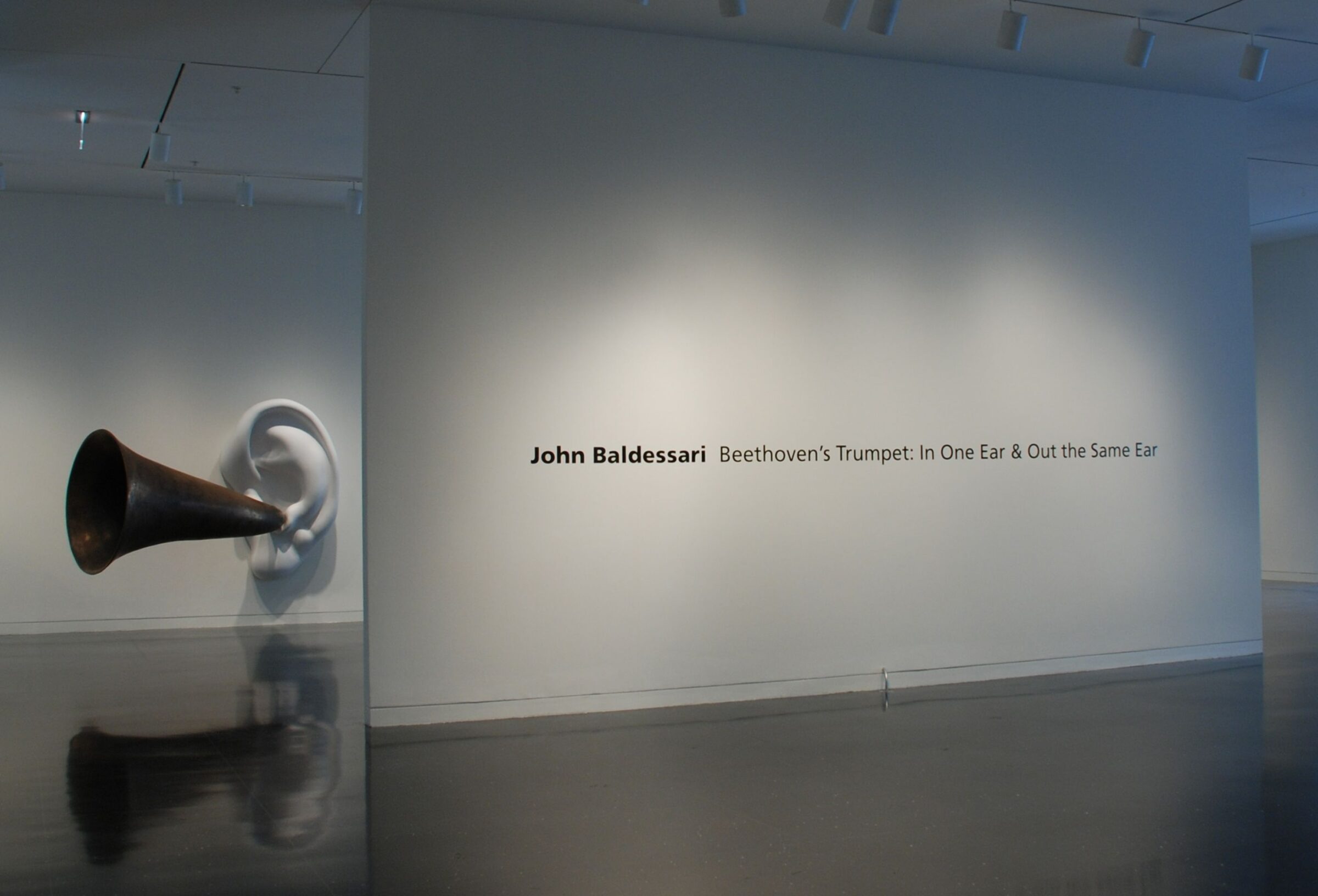 A freestanding gallery wall with black text that reads "John Baldessari: Beethoven's Trumpet: In One Ear & Out the Same Ear" with a sculpture of a massive white ear with a large brass ear horn sticking out of it.
