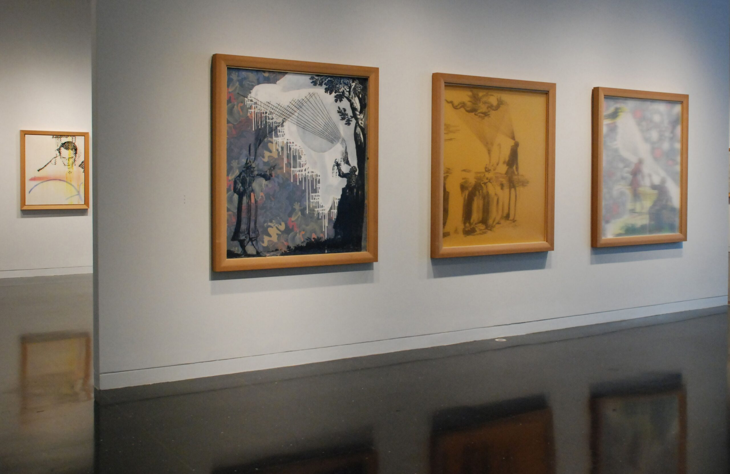 A freestanding, white gallery wall with three collages in wood frames installed in a row. Another framed image is visible in the gallery behind the wall.