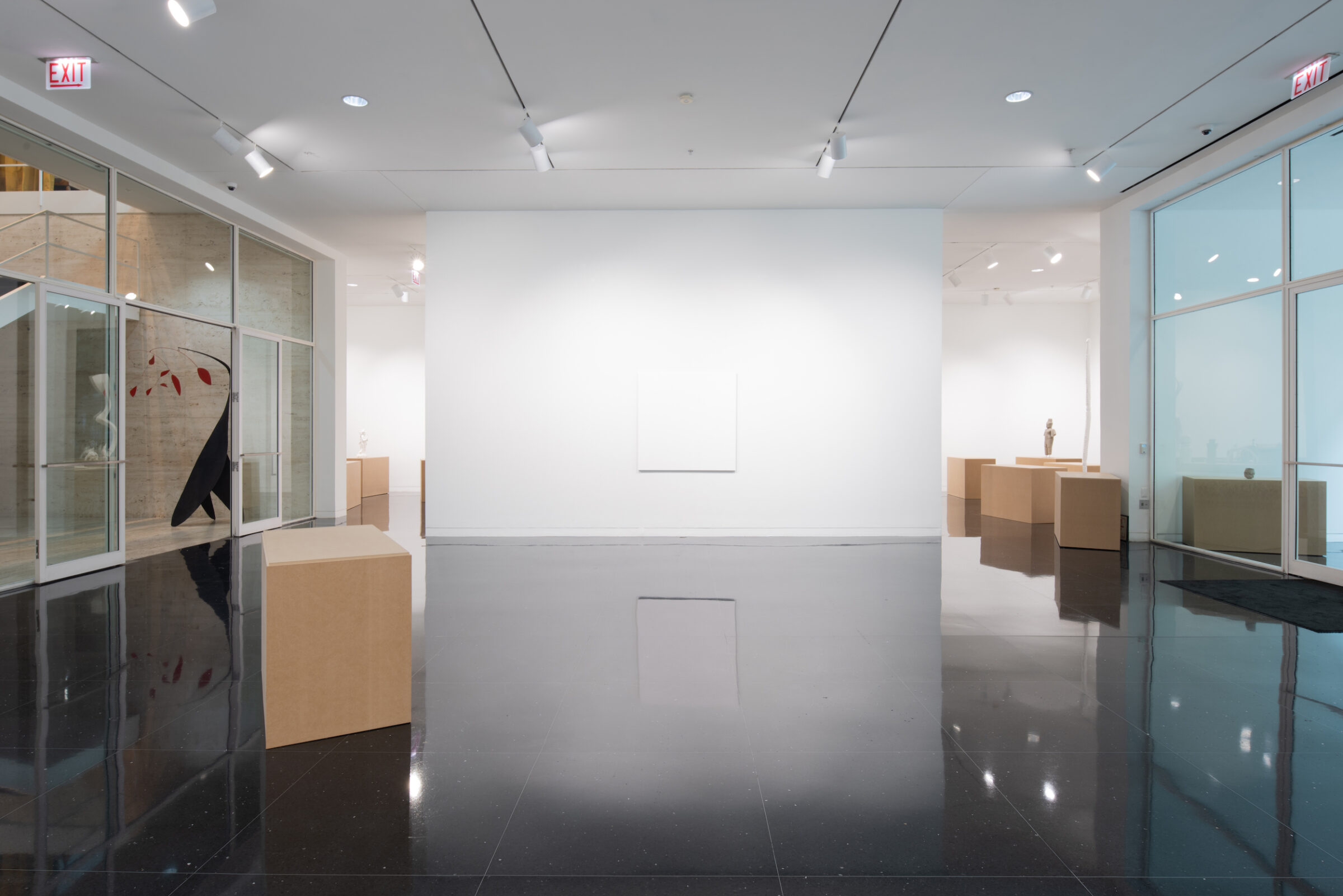 A white walled gallery containing several pedestals resembling cardboard boxes. One on freestanding wall, a large square canvas the identical color of the wall hangs centered. In a far gallery, a sculpture of a standing figure is visible.