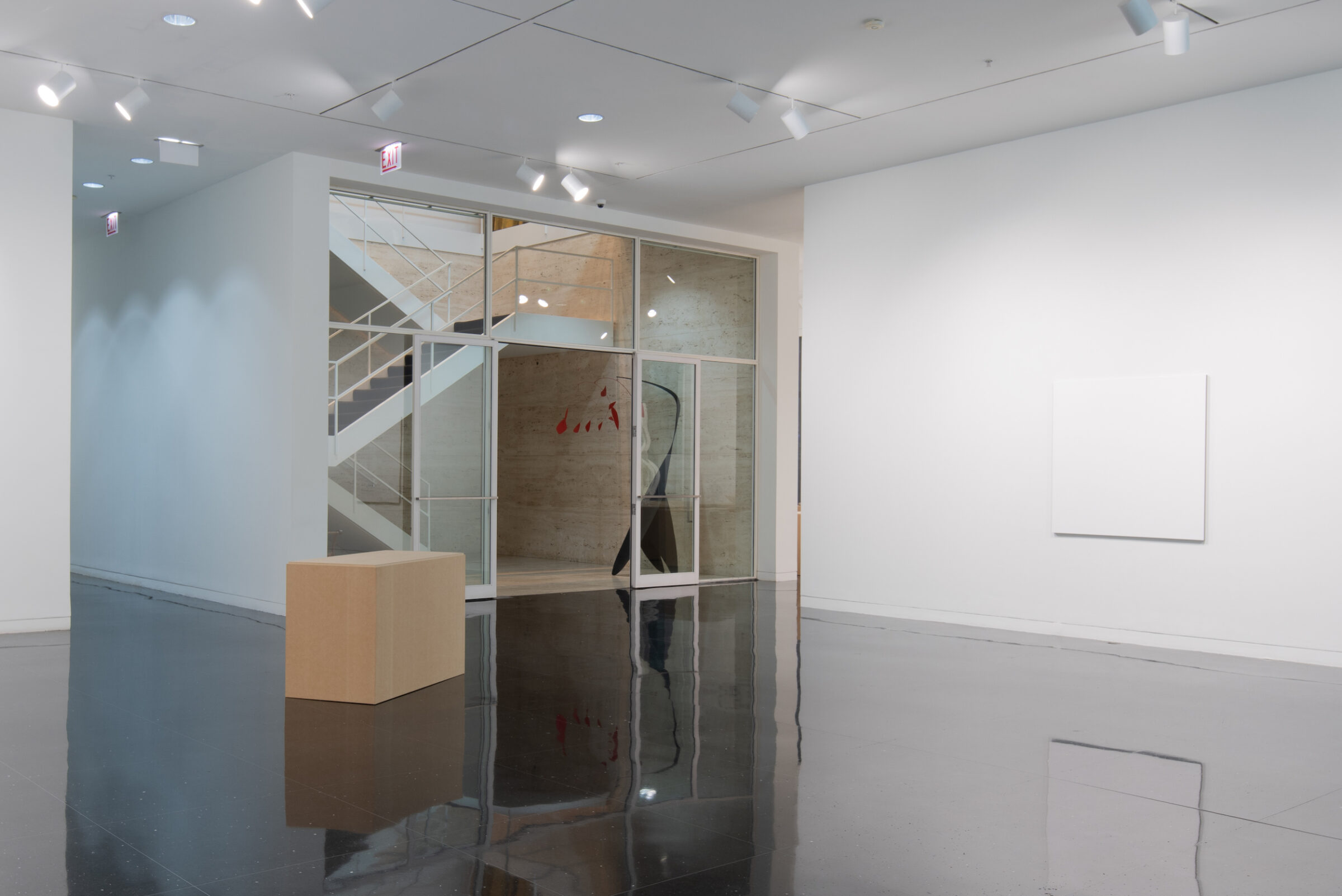 A white walled gallery with one pure white square canvas installed on a freestanding wall of identical color and one pedestal resembling a cardboard box. In the center of these two works, the Mies van der Rohe stairwell is visible.