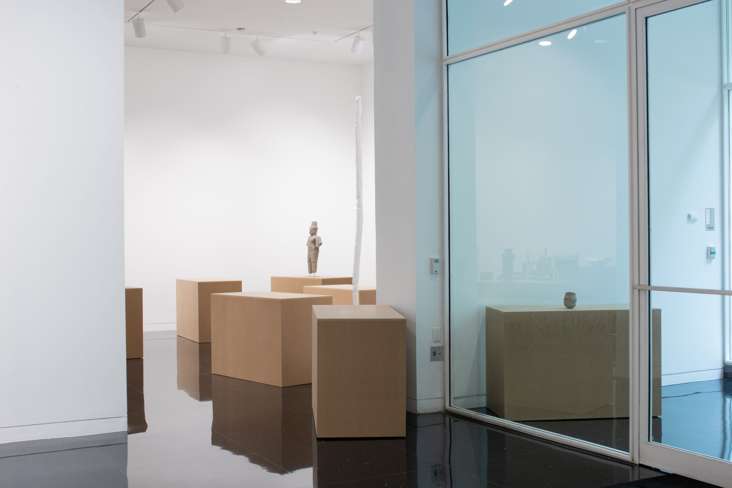 Several pedestals resembling cardboard boxes in a white walled gallery. In the back of the gallery, an armless, gray standing sculpture of a figure stands atop a pedestal.