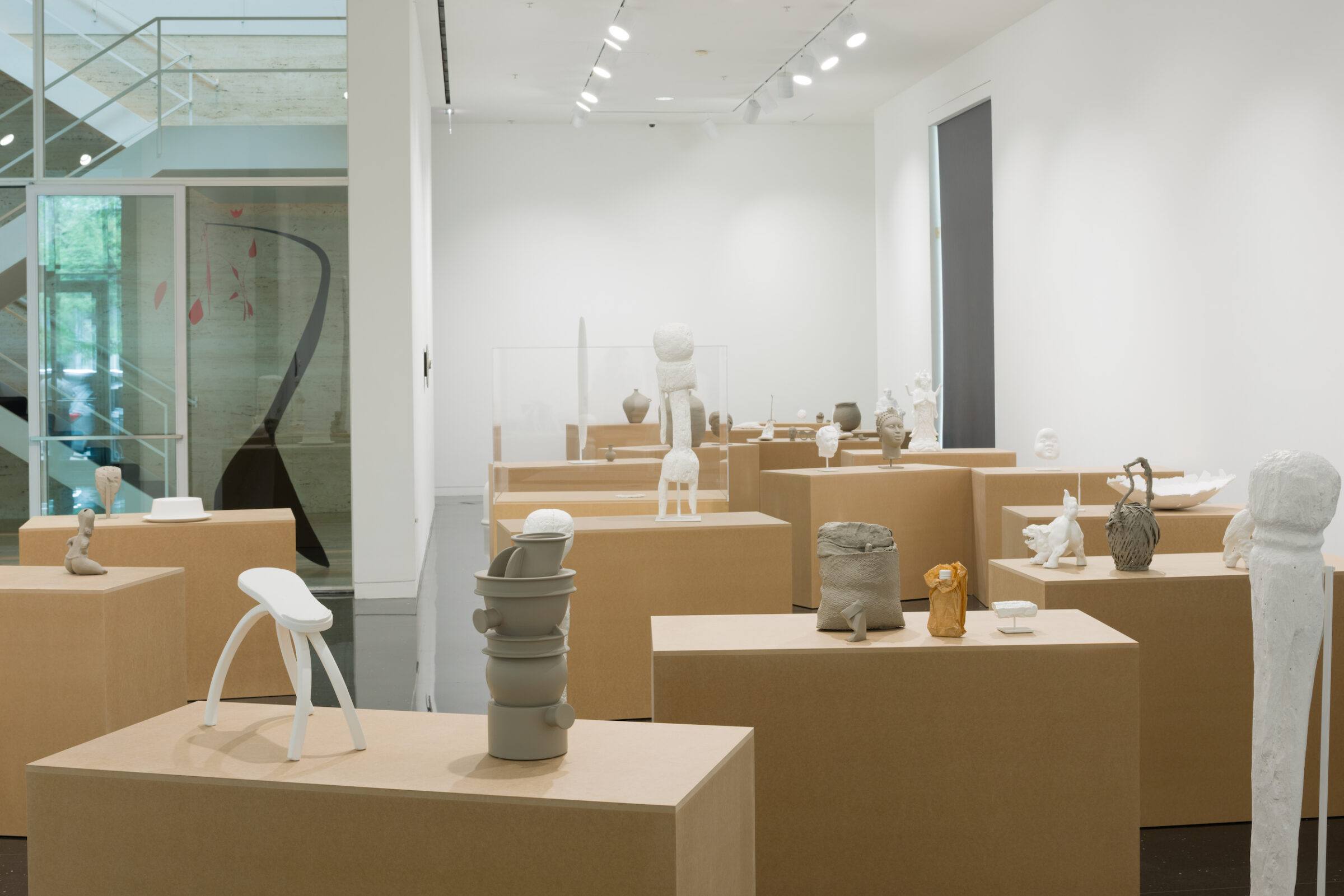 Over a dozen pedestals resembling cardboard boxes installed in a white gallery. Atop each pedestal are several unglazed ceramic sculptures, some fired and some unfired, raw clay. Sculptures include heads and busts, vases, baskets, and animals.